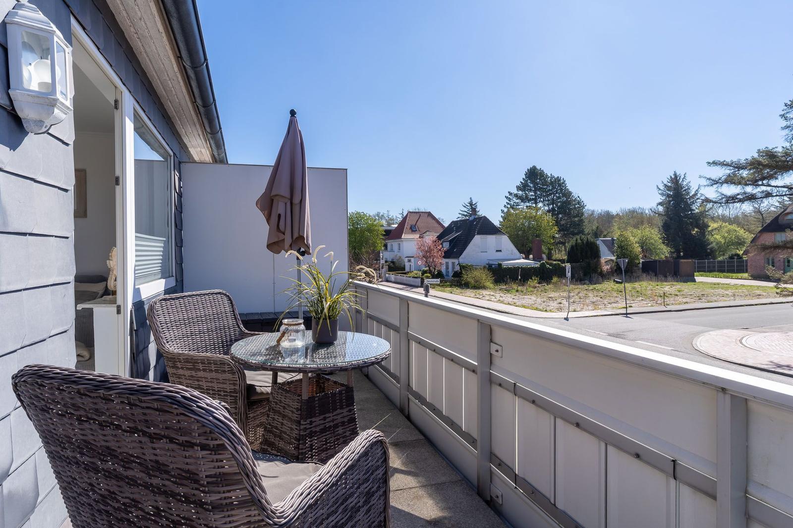 Terrasse mit Tisch, zwei Stühlen und geschlossenem Sonnenschirm. Blick auf Straße und Wohnhäuser.
