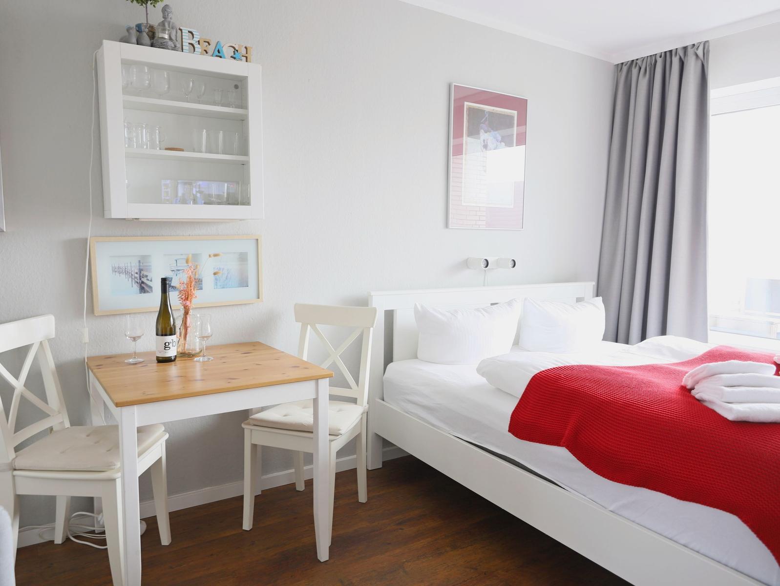 Ein Schlafzimmer mit weißem Bett, rotem Deckenbezug, Esstisch und Glaswand.