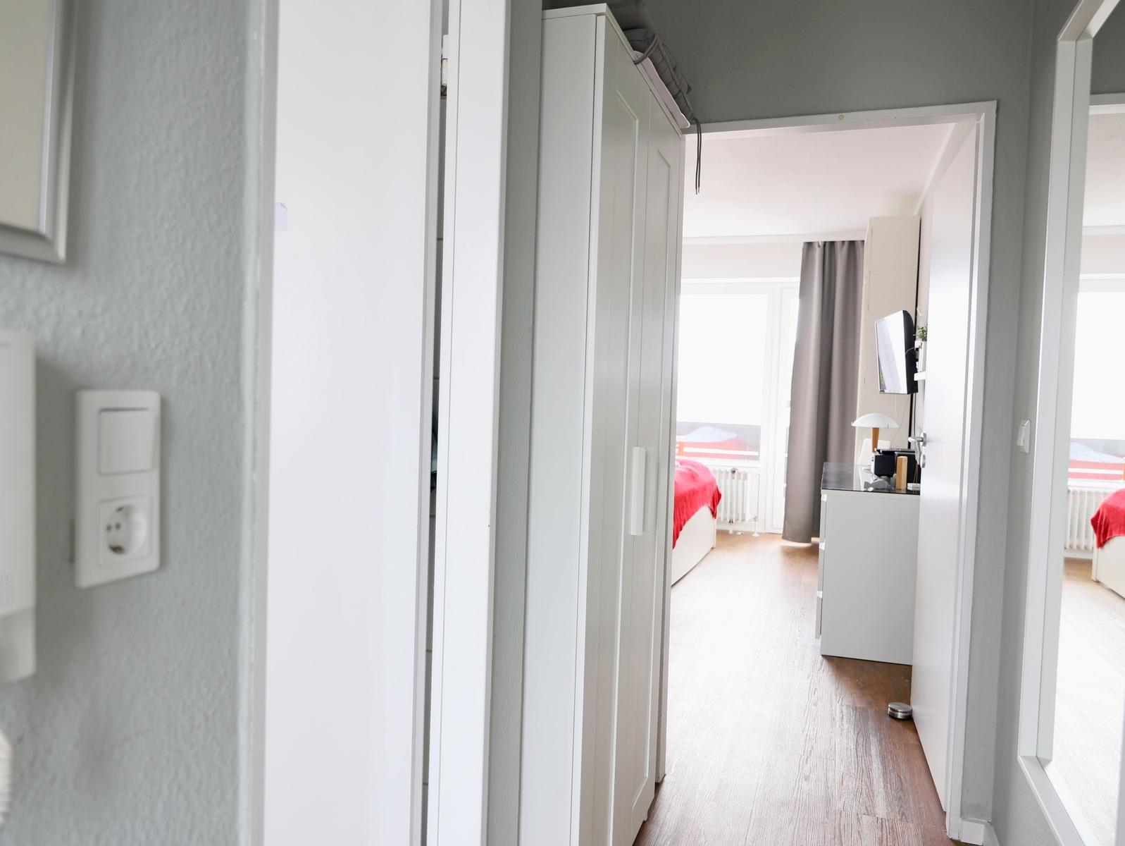 Schlafzimmer mit weißem Schrank und rotem Bett. Blick auf Fenster mit Vorhängen.