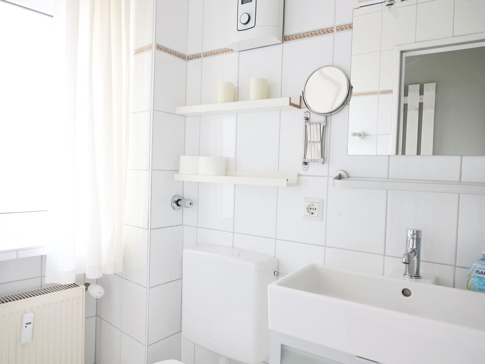 Badezimmer mit Toilette, Waschbecken und Spiegel. Weiße Fliesen und Regale.