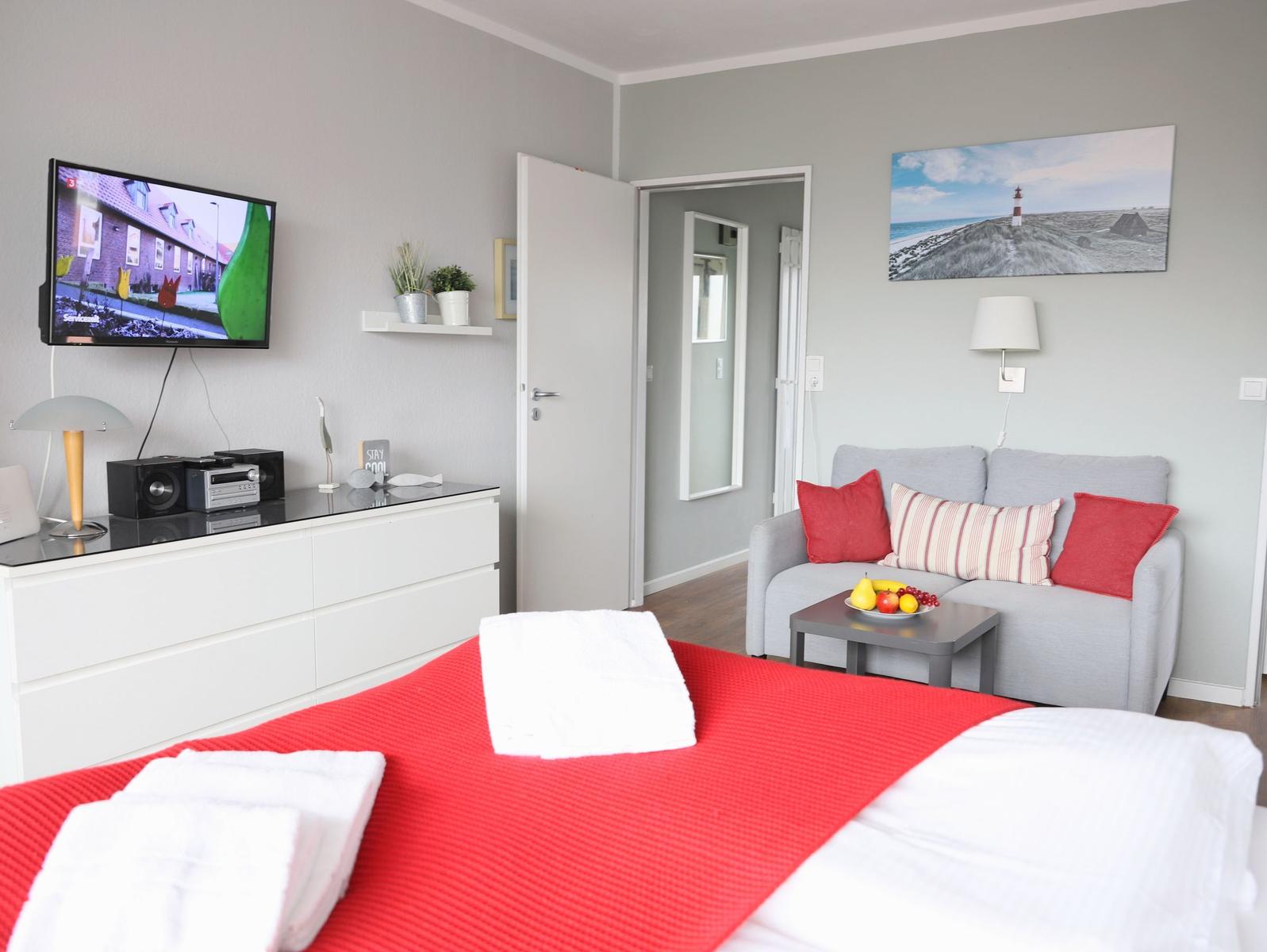 Schlafzimmer mit Bett, Sofa, Fernseher und Wandbild. Tisch mit Obst und roten Kissen.