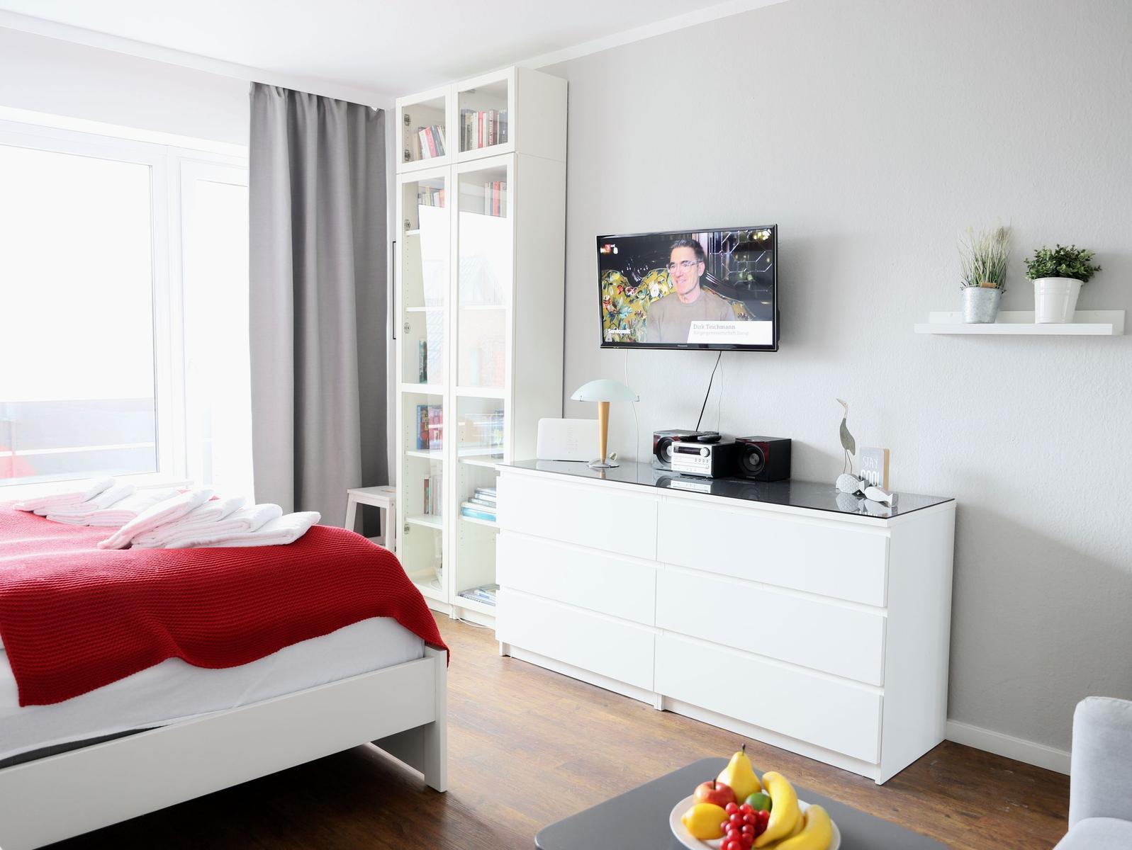 Schlafzimmer mit Bett, Fernseher und Bücherregal. Auf dem Tisch Obst.