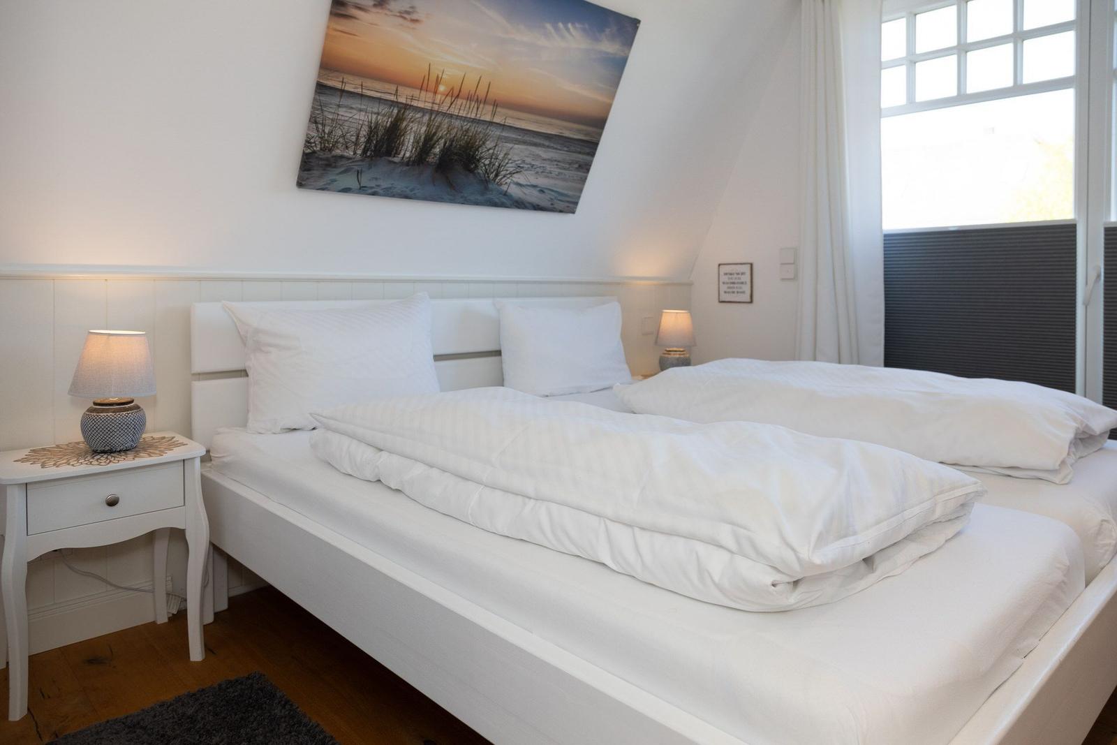 Schlafzimmer mit weißem Bett, Nachttisch und Wandbild. Fenster mit Jalousie.