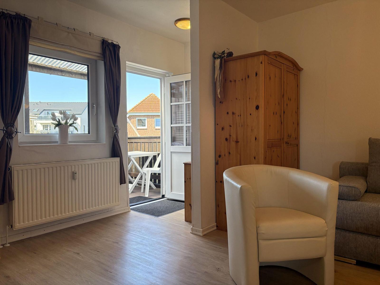 Wohnraum mit Sitzgelegenheit, Holzschrank und Blick auf Balkon mit Tisch und Stühlen.