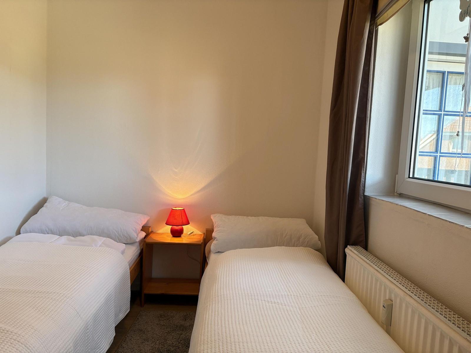 Zwei Einzelbetten mit weißen Bettwäschen, Nachttisch mit Lampe und Fenster mit Vorhang.