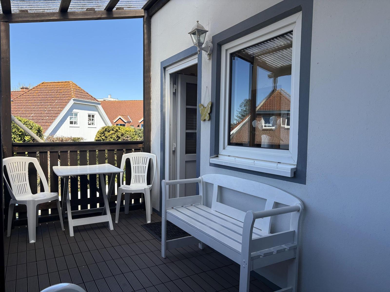 Terrasse mit weißem Tisch, zwei Stühlen und Bank. Blick auf Haus mit rotem Dach.