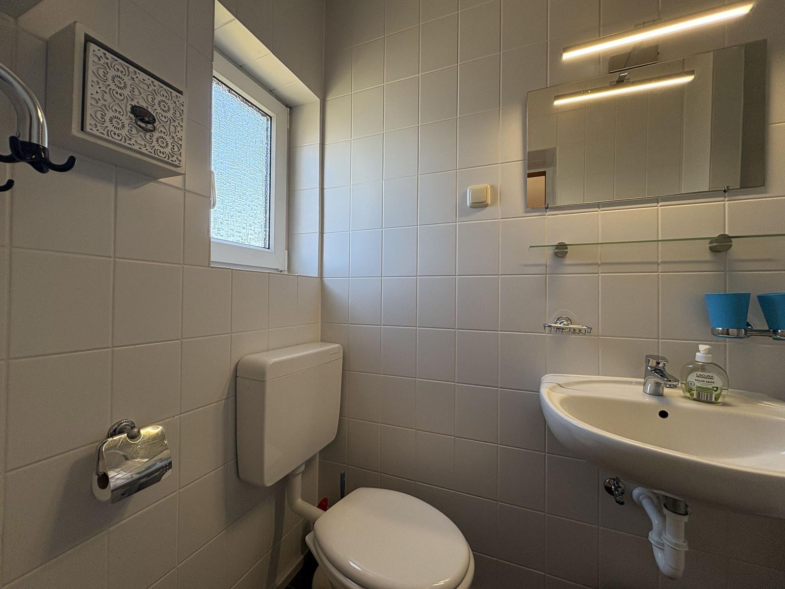 Badezimmer mit Toilette, Waschbecken und Spiegel. Fenster mit Gitter und Toilettenpapierhalter.