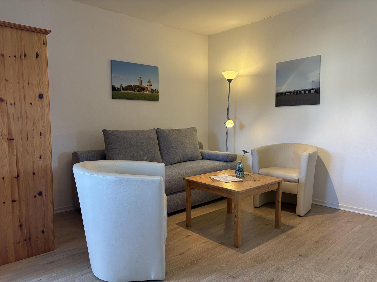 Wohnzimmer mit Sofa, zwei Sesseln und Holztisch. An den Wänden hängen Bilder.