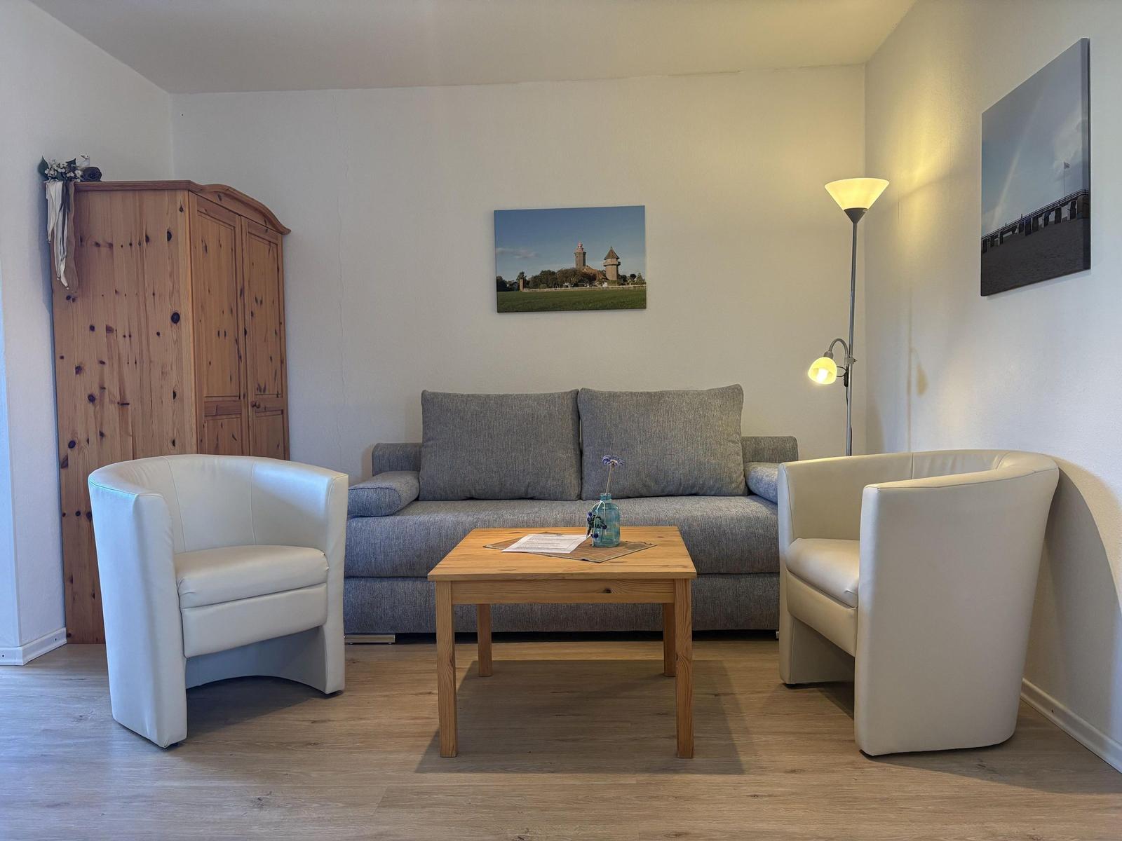 Wohnzimmer mit Sofa, zwei weißen Sesseln und Holztisch. An der Wand hängen Bilder.