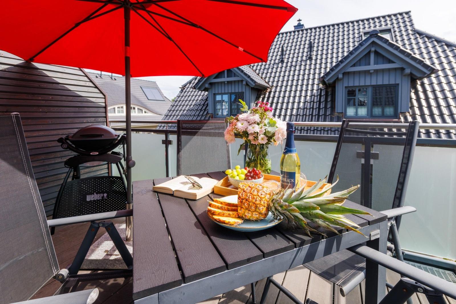 Terrasse mit Tisch, Stühlen, Sonnenschirm und Grill. Obst, Wein und Blumen sind darauf platziert.