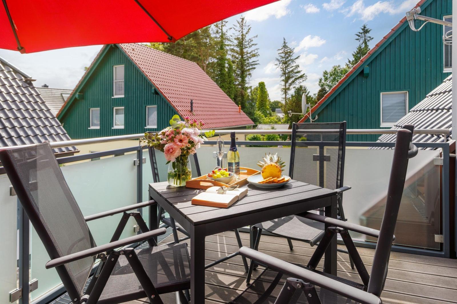 Terrasse mit Tisch, Stühlen und rotem Sonnenschirm. Im Hintergrund grüne Häuser und Bäume.