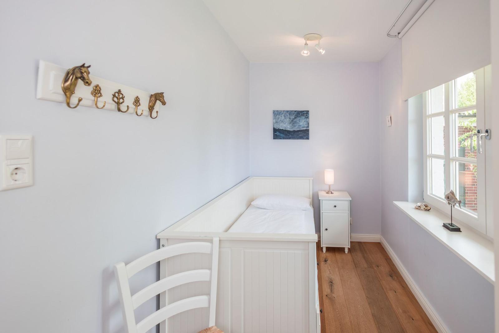 Schlafzimmer mit weißem Bett, Nachttisch und Fenster. An der Wand hängt ein Bild und ein Hakenbrett mit Pferdekopf.