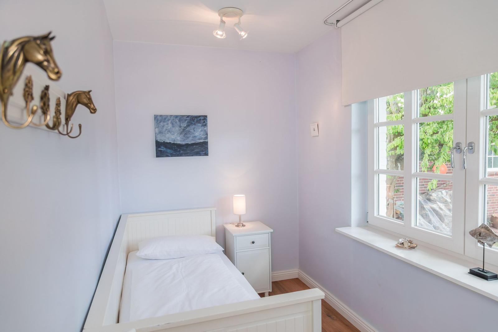 Schlafzimmer mit weißem Bett, Nachttisch und Fenster mit Blick auf Bäume.