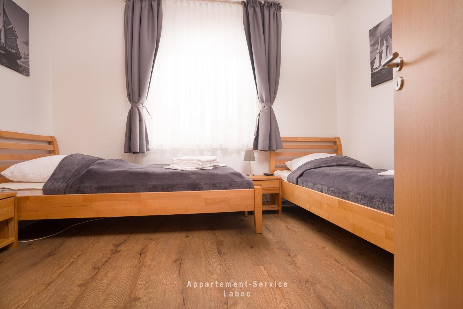Zwei Einzelbetten mit grauer Bettwäsche, Holzboden und Fenster mit grauen Vorhängen.
