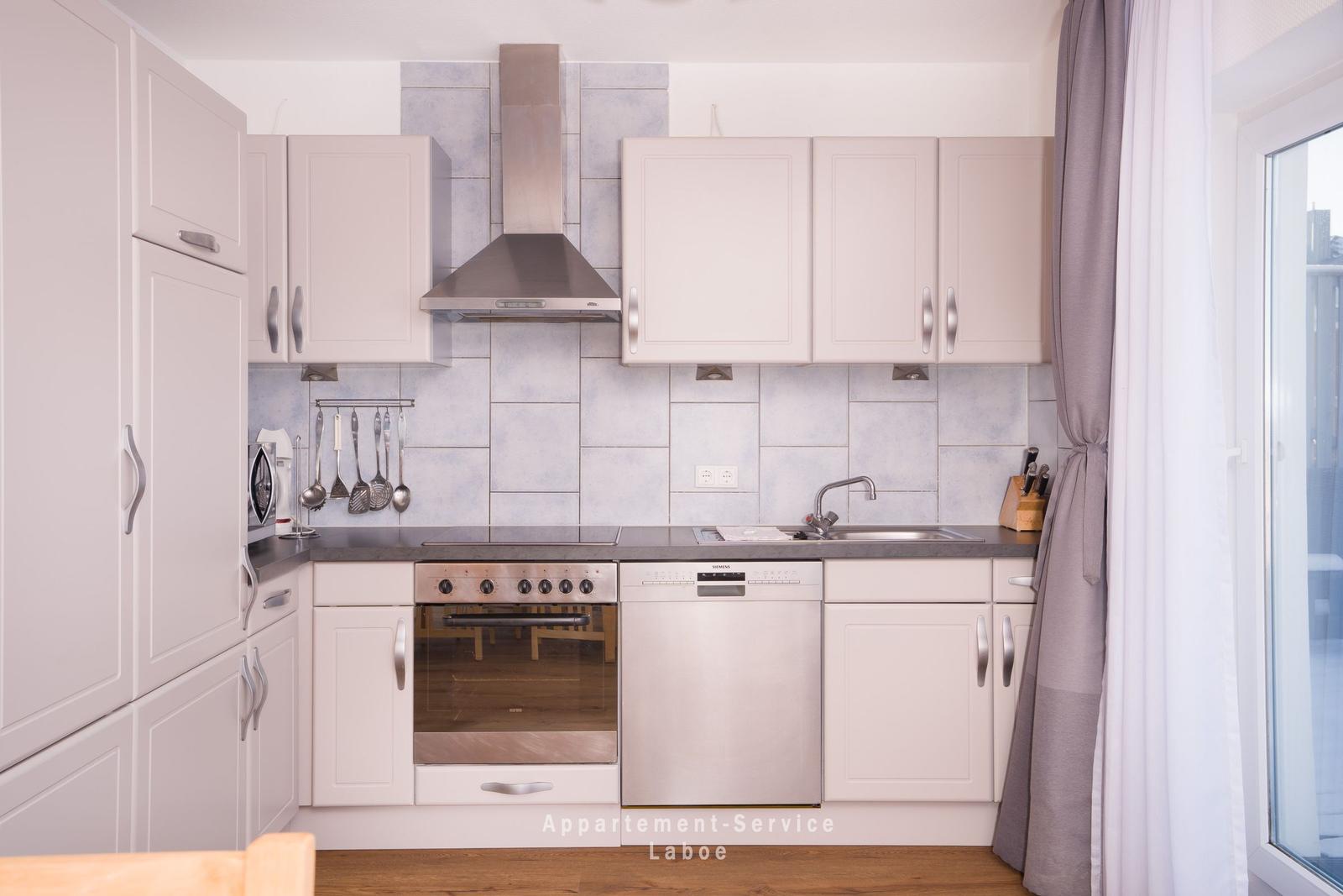 Küche mit weißen Schränken, Edelstahlspüle und Herd. Fenster mit Vorhängen.