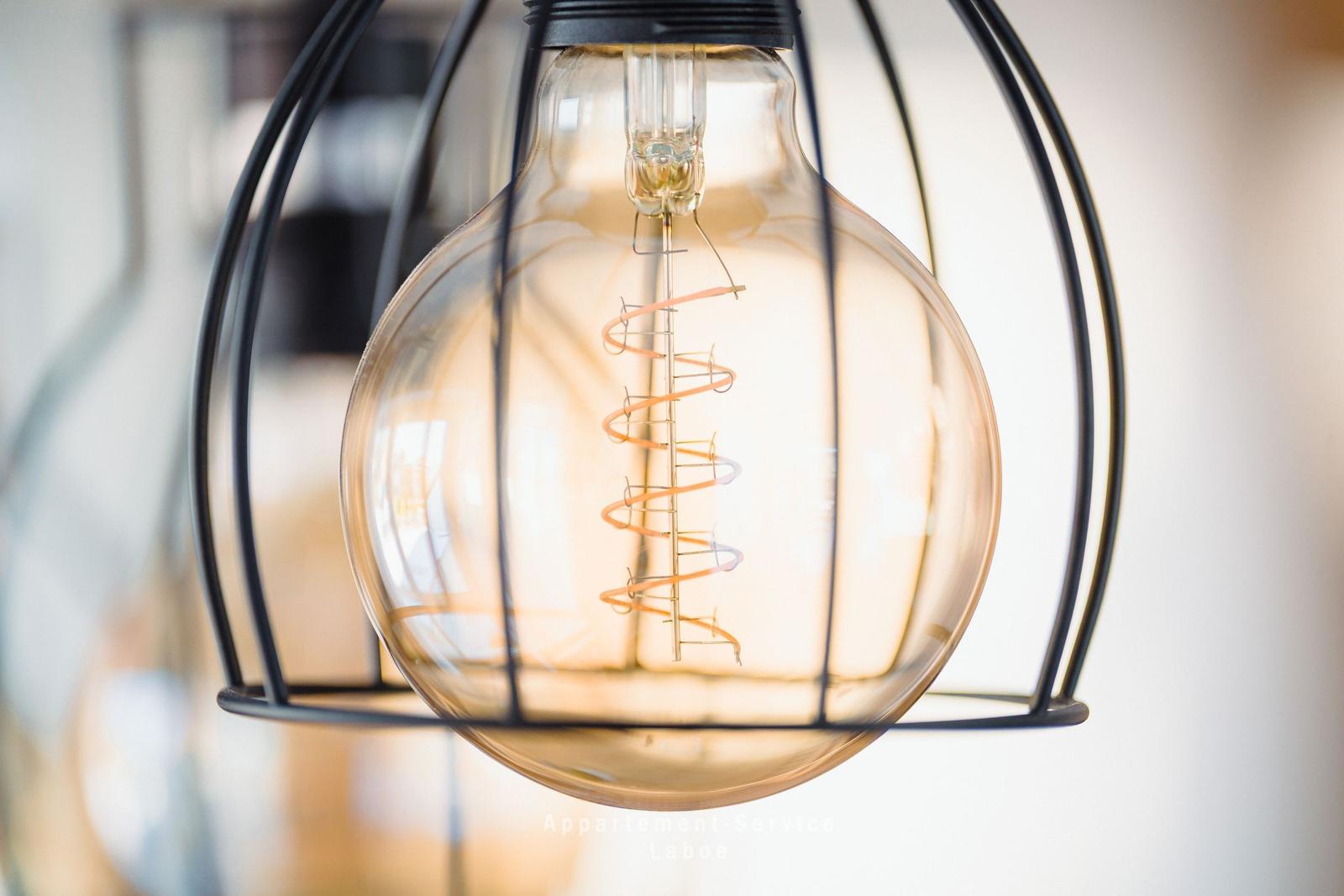 Eine gläserne Glühlampe mit Spiralfilament in einem schwarzen Metallkäfig hängt an der Decke.