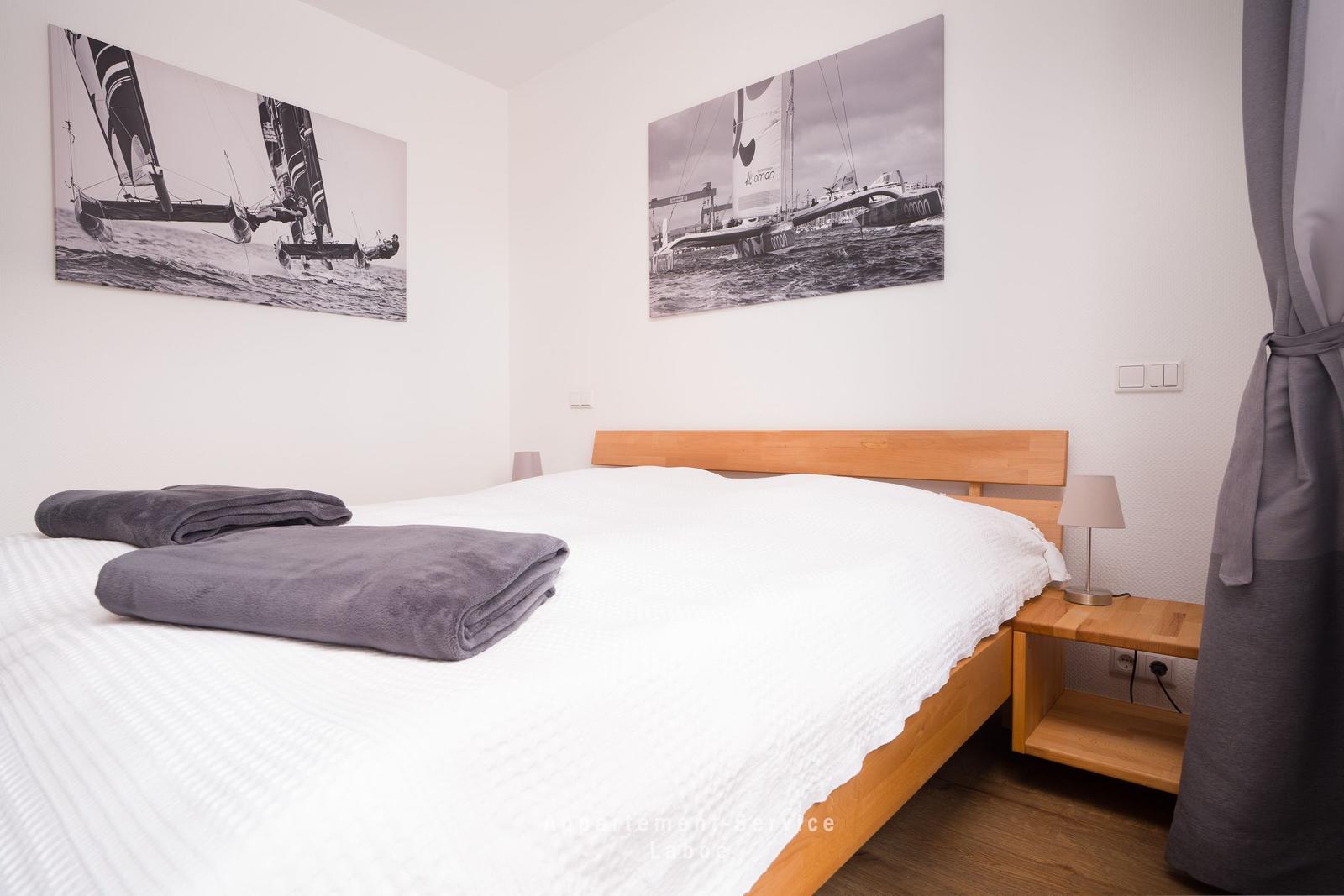 Doppelbett mit weißen Laken und grauen Decken. Wandbilder zeigen Segelboote.