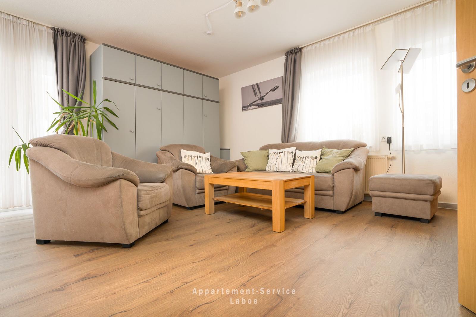 Wohnzimmer mit Sofa, Sesseln und Holztisch. Holzdielenboden und Fenster mit Vorhängen.