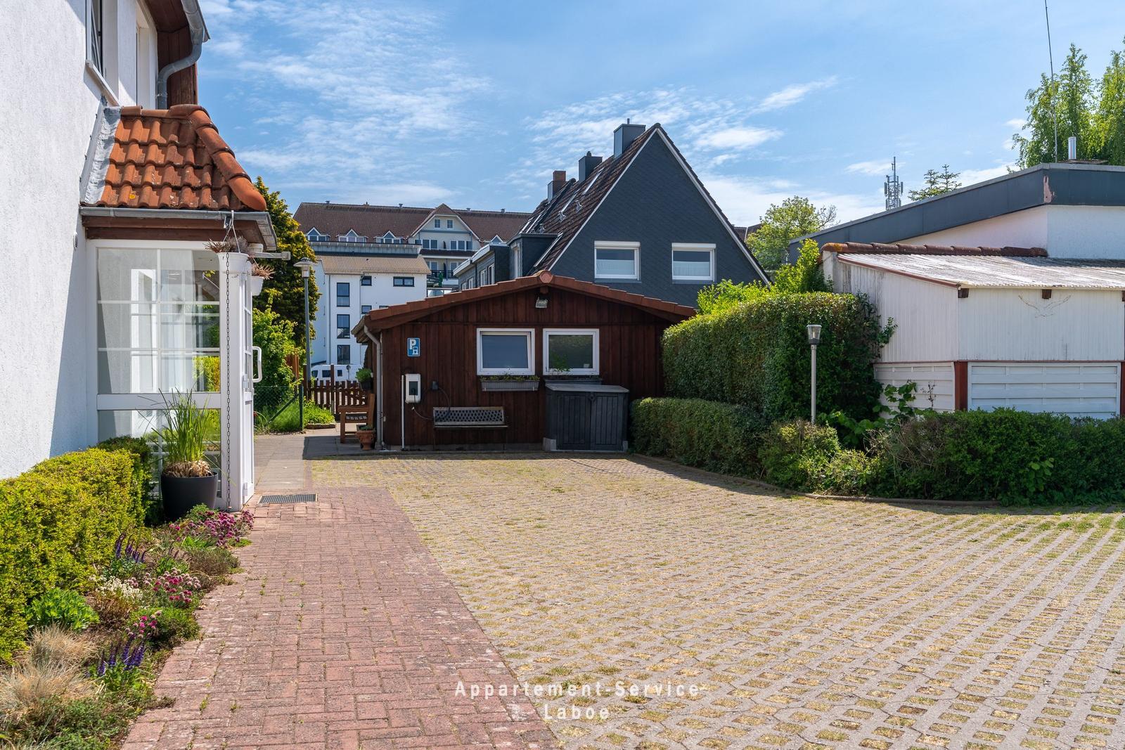 Hinterhof mit gepflastertem Platz, kleinem Holzhaus und grüner Hecke.