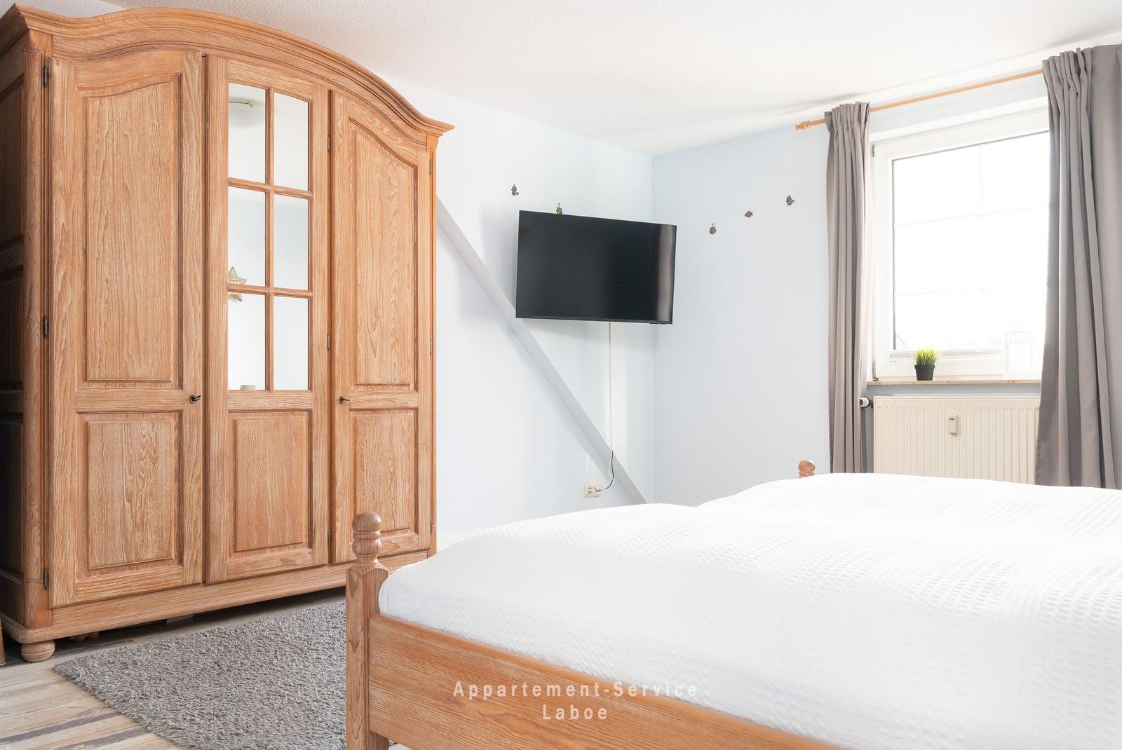 Schlafzimmer mit Holzbett, Kleiderschrank und Wandfernseher. Fenster mit grauen Vorhängen.