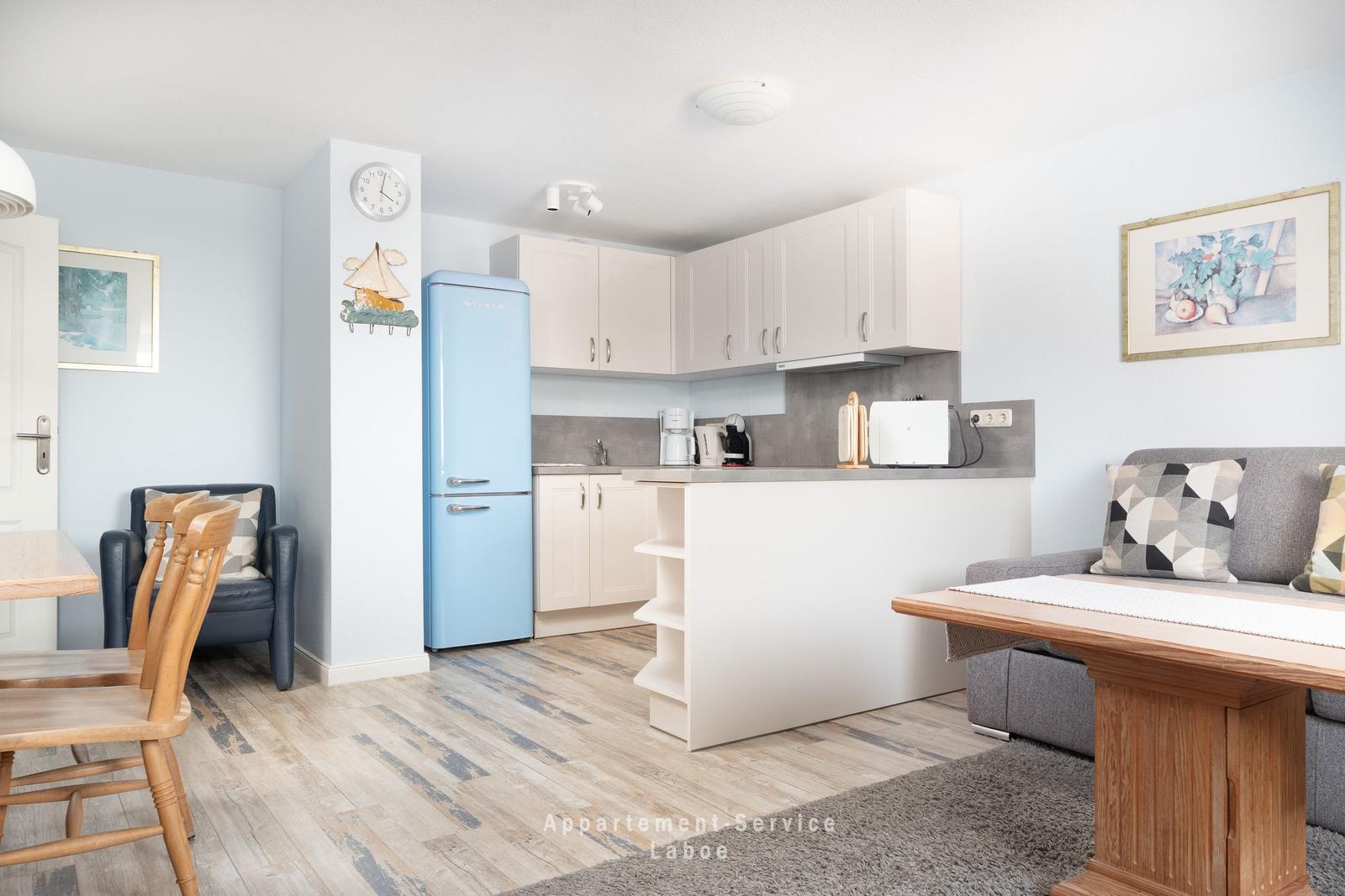 Küche mit hellen Schränken und blauem Kühlschrank. Wohnbereich mit Sofa und Holztisch.