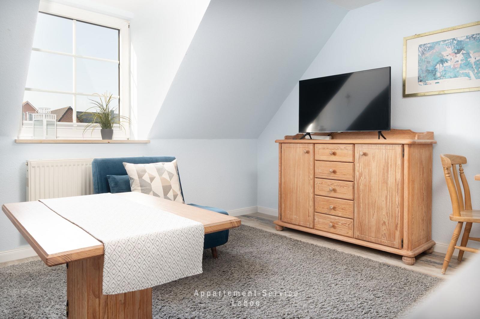 Ein Raum mit Dachschräge, TV-Schrank, Tisch und Sessel. Fenster mit Pflanze.