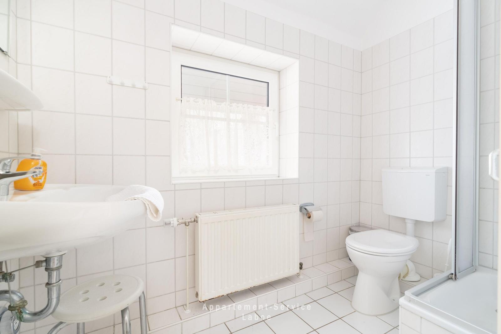 Badezimmer mit Toilette, Waschbecken und Dusche. Weiße Fliesen und Fenster mit Vorhang.