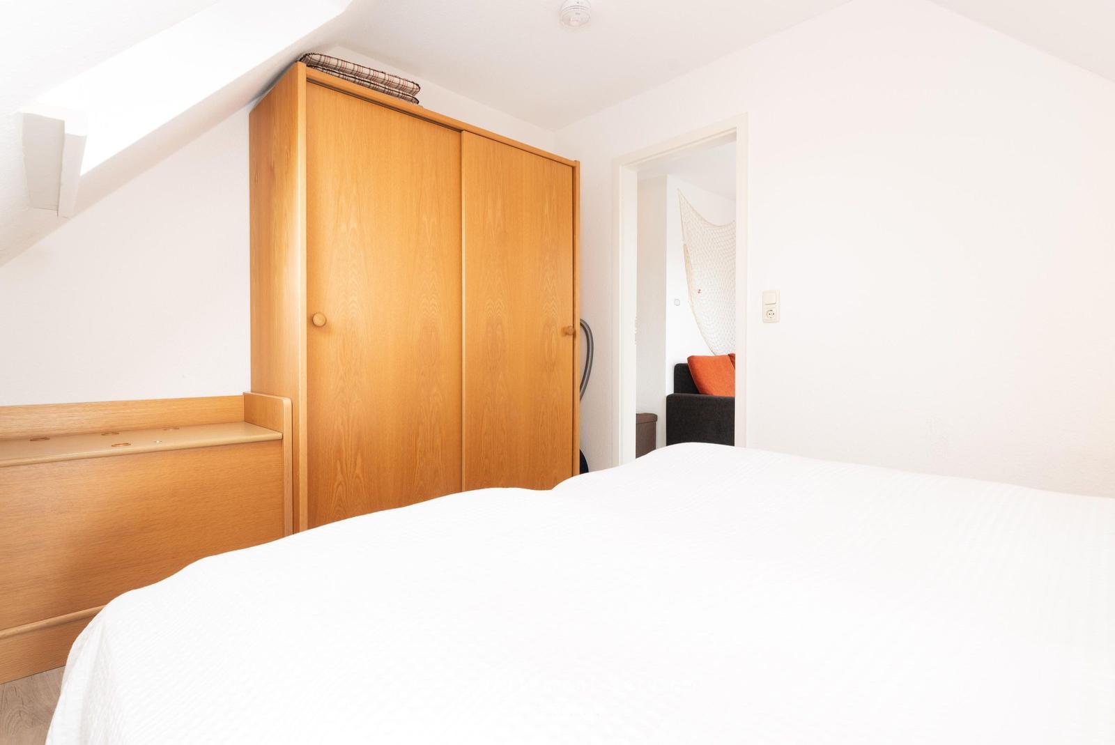 Schlafzimmer mit Dachschräge, Holzschrank und Bett. Durchgang zu Wohnbereich mit Sofa.