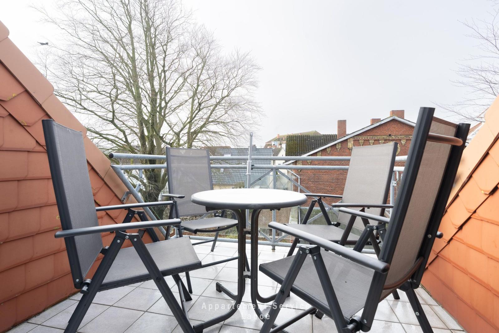 Terrasse mit Tisch und vier Stühlen, umgeben von roten Ziegeldächern und Bäumen.