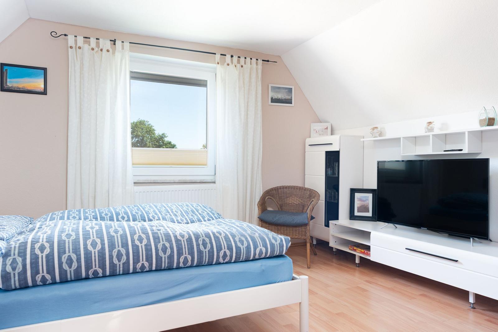 Schlafzimmer mit Bett, Fenster, TV und Stuhl.