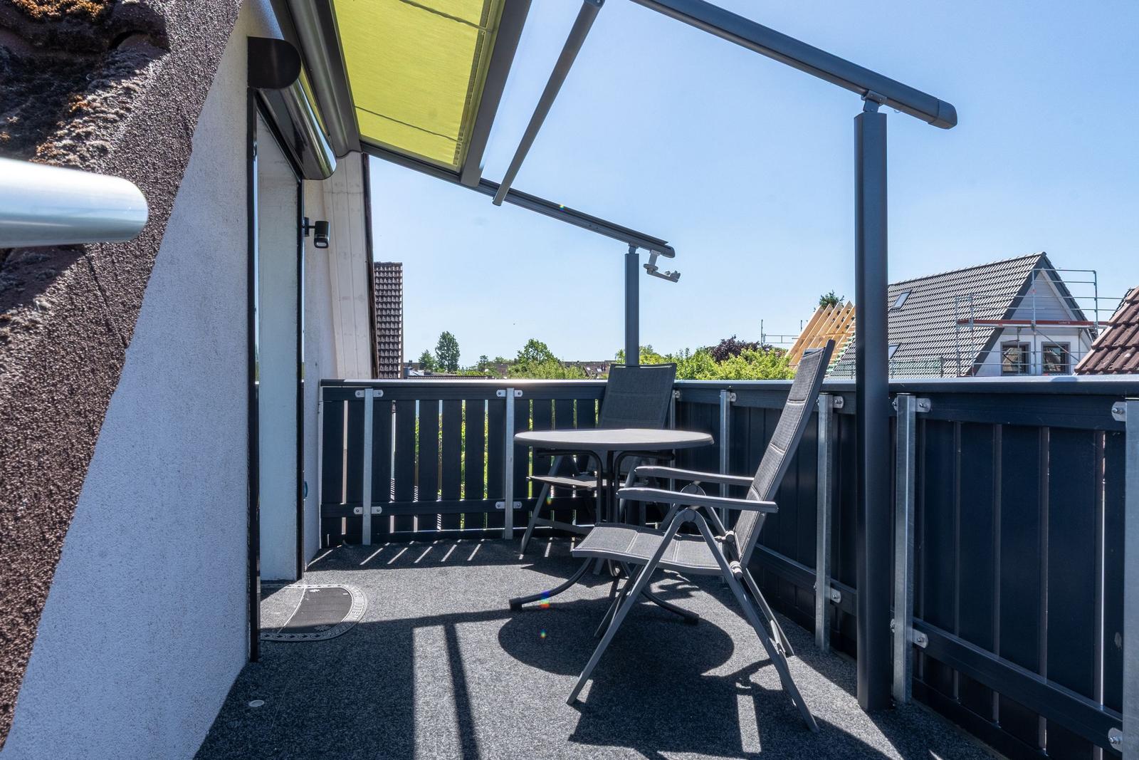 Terrasse mit Tisch, zwei Stühlen und gelbem Sonnenschutz. Blick auf Wohnhäuser und Bäume.