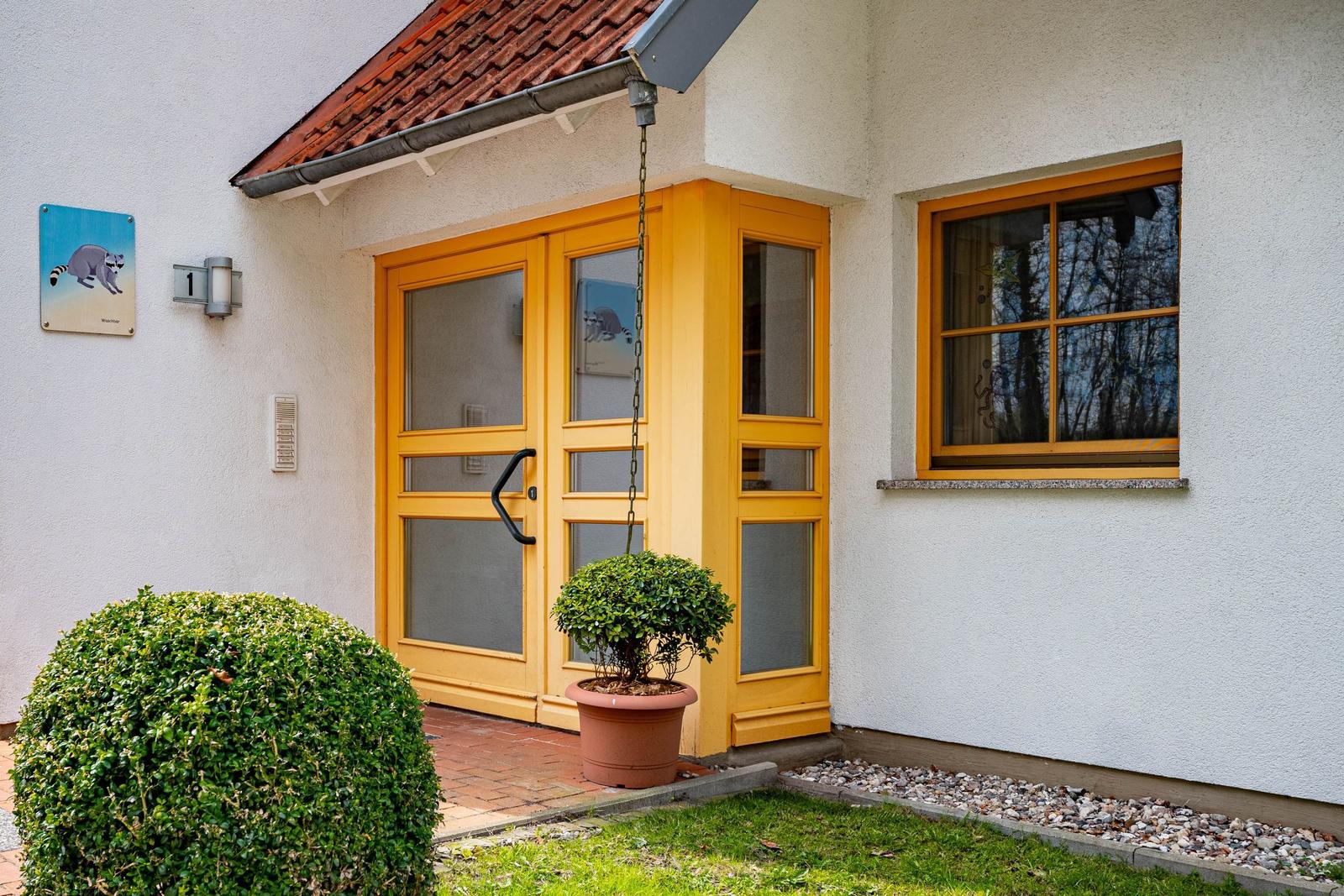 Eingangstür mit gelbem Rahmen und Pflanzen vor einem Haus mit weißen Wänden.