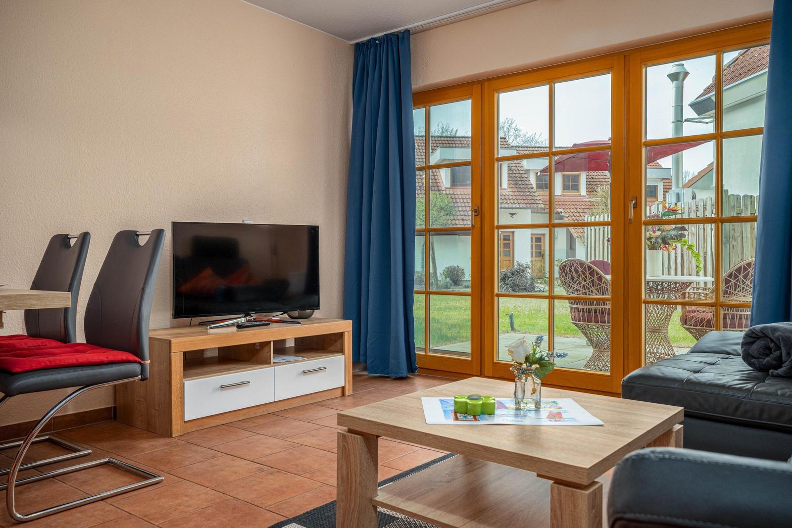 Wohnzimmer mit TV, Tisch und Stühlen, Blick auf Garten durch Glastüren.