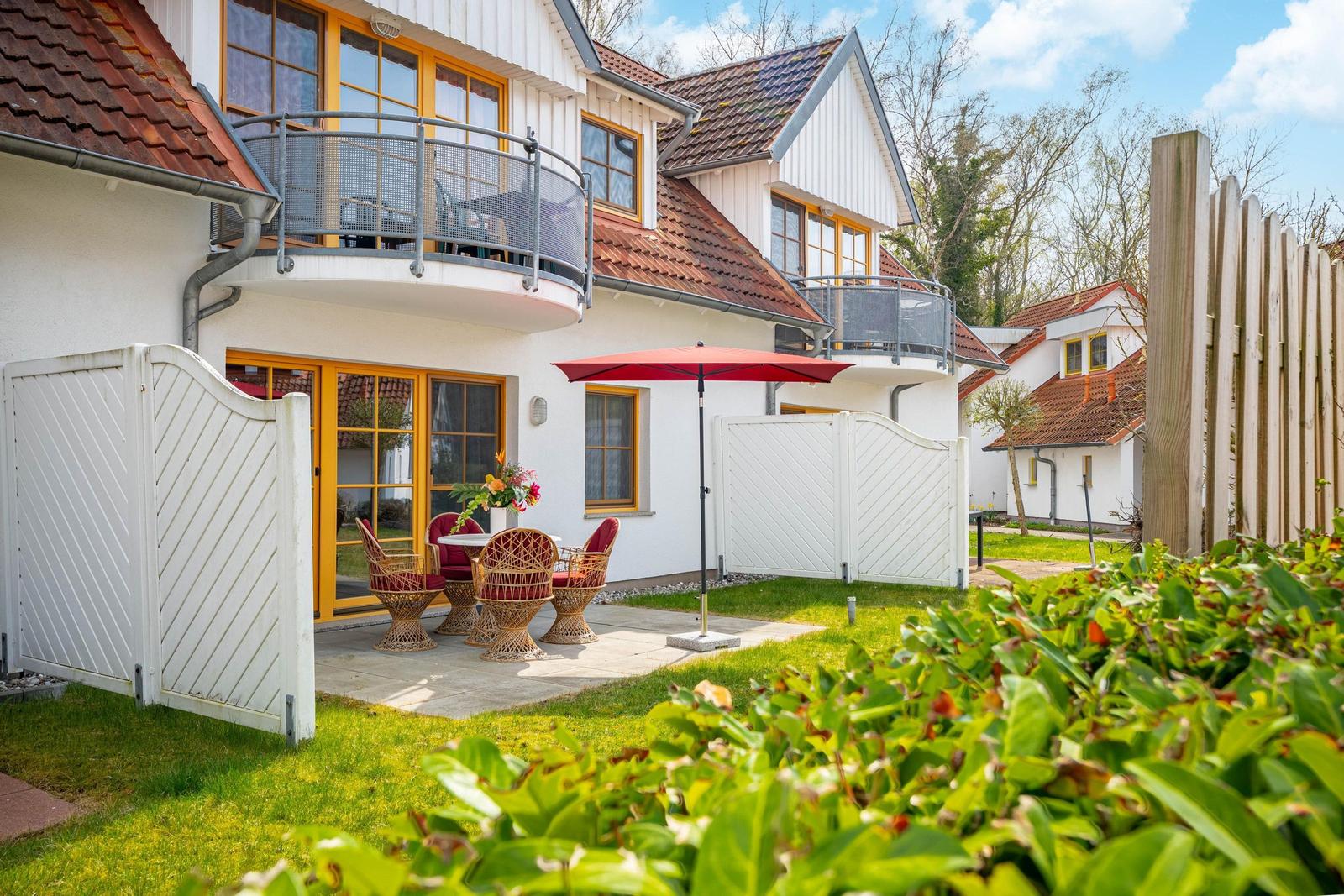 Haus mit Terrasse, Garten und rotem Sonnenschirm. Weiße Wände, gelbe Fenster und rote Dachziegel.