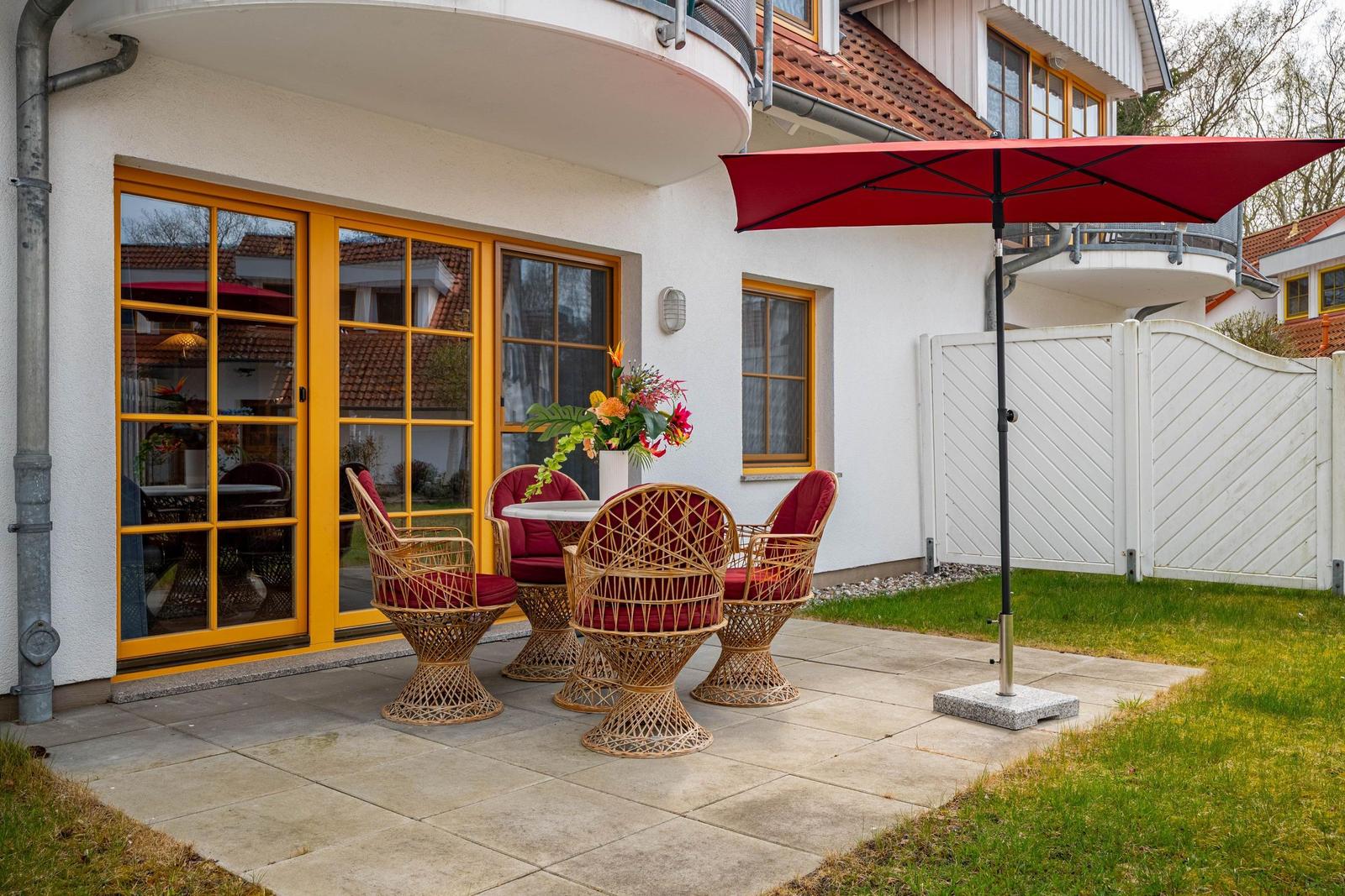 Terrasse mit rotem Sonnenschirm und Rattanmöbeln vor weißem Haus mit gelben Fenstern.