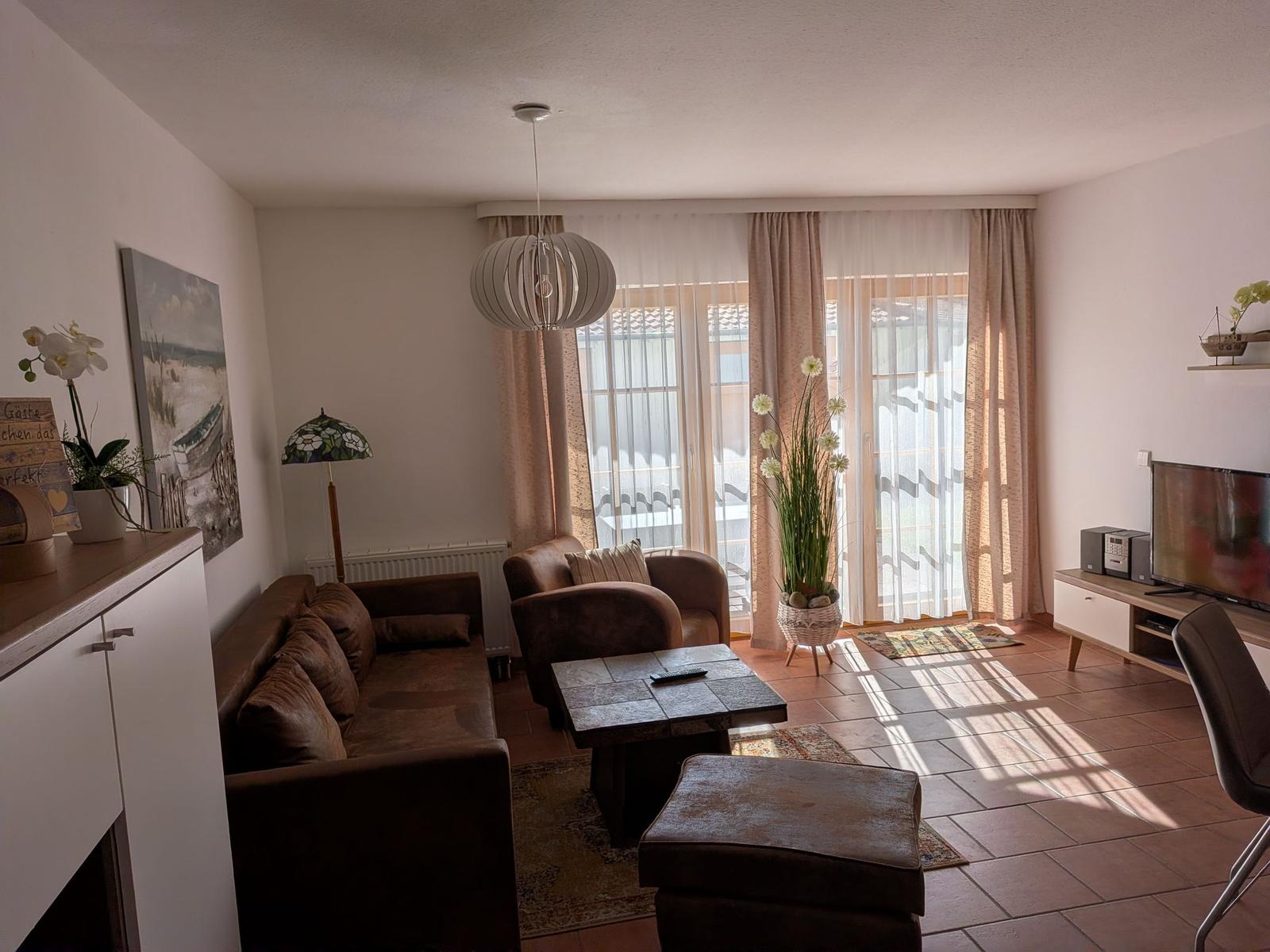 Wohnzimmer mit Sofa, Sessel und TV. Durchgang zur Terrasse mit Gardinen.