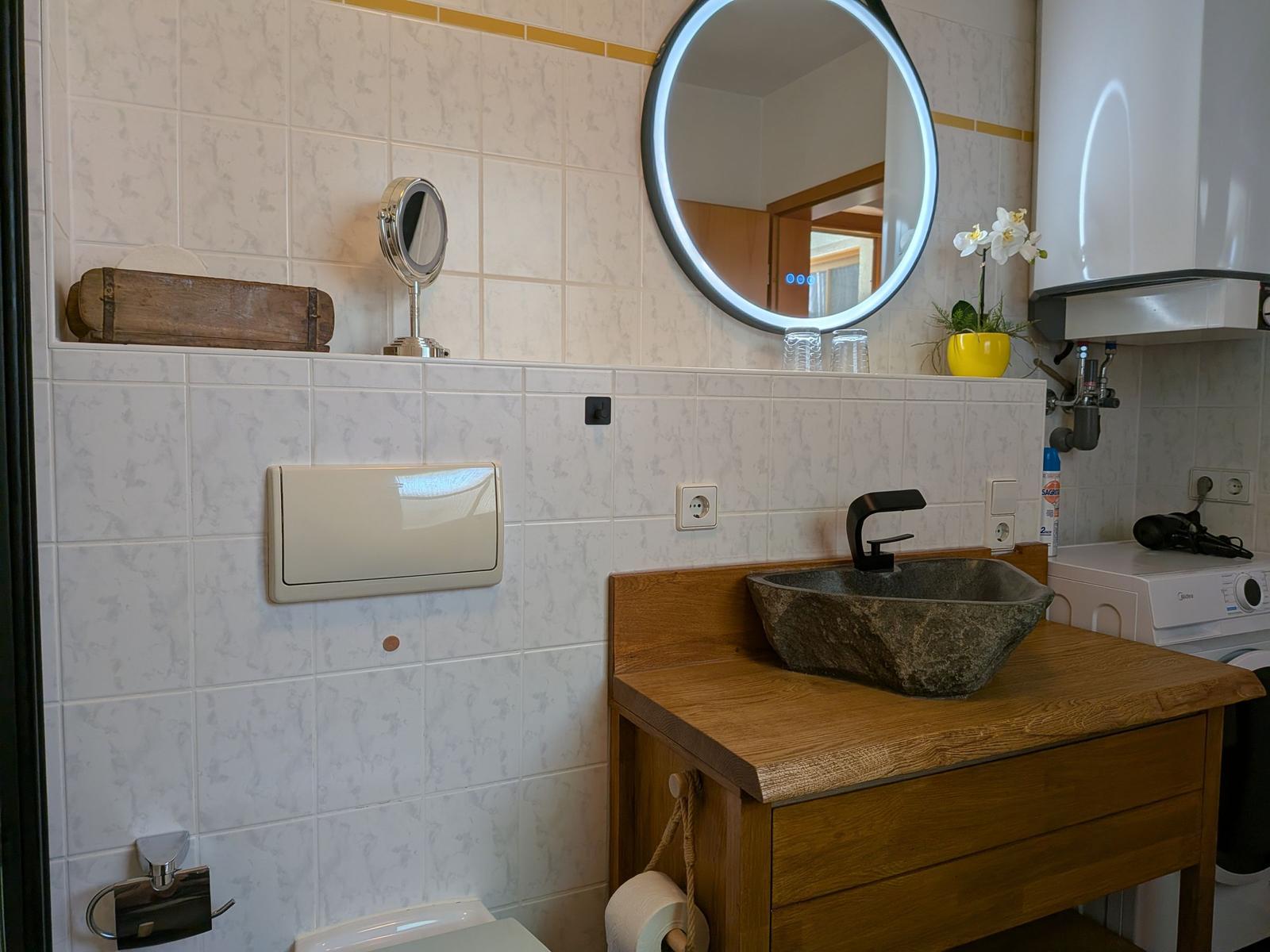 Bad mit Steinwaschbecken, Holzwaschtisch und runden Spiegel mit LED-Rand.