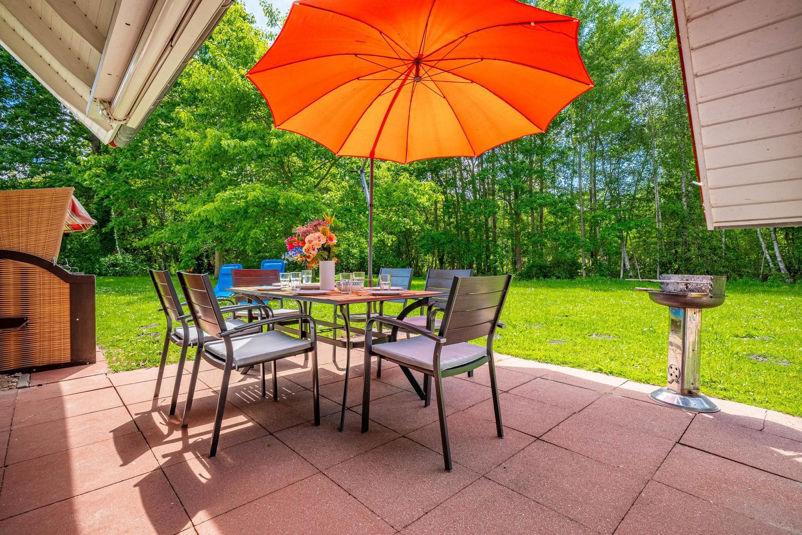 Terrasse mit Tisch, Stühlen und orangefarbenem Sonnenschirm. Grill und Garten im Hintergrund.