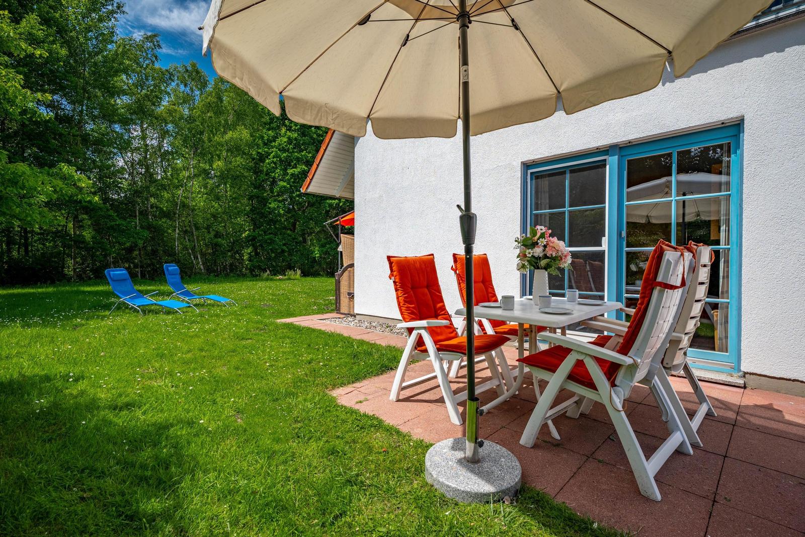 Terrasse mit Tisch, Stühlen und Sonnenschirm neben weißer Wand mit blauen Fenstern.