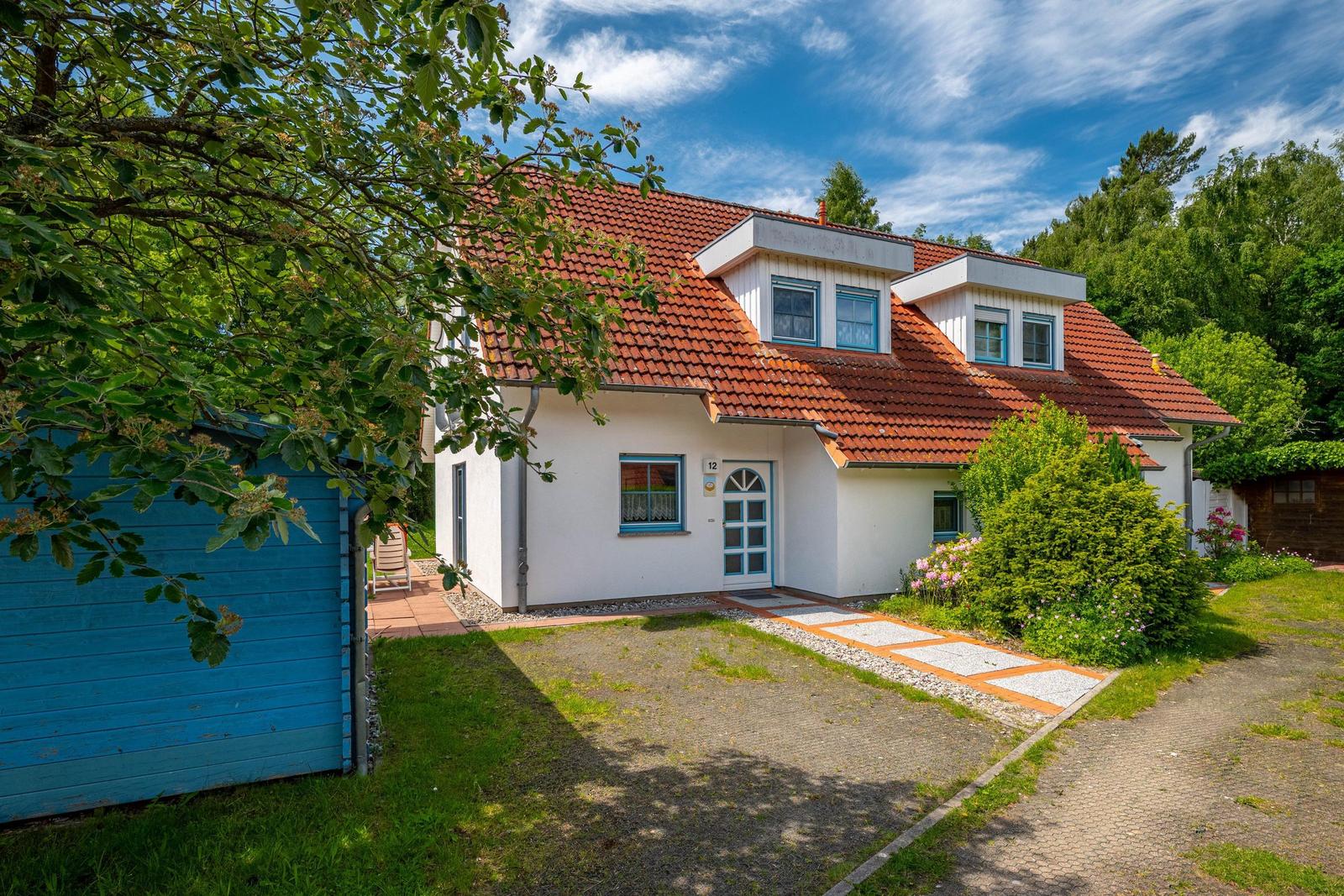 Ein weißes Haus mit rotem Dach und blauen Fensterrahmen. Vorhof mit Weg und Grünflächen.