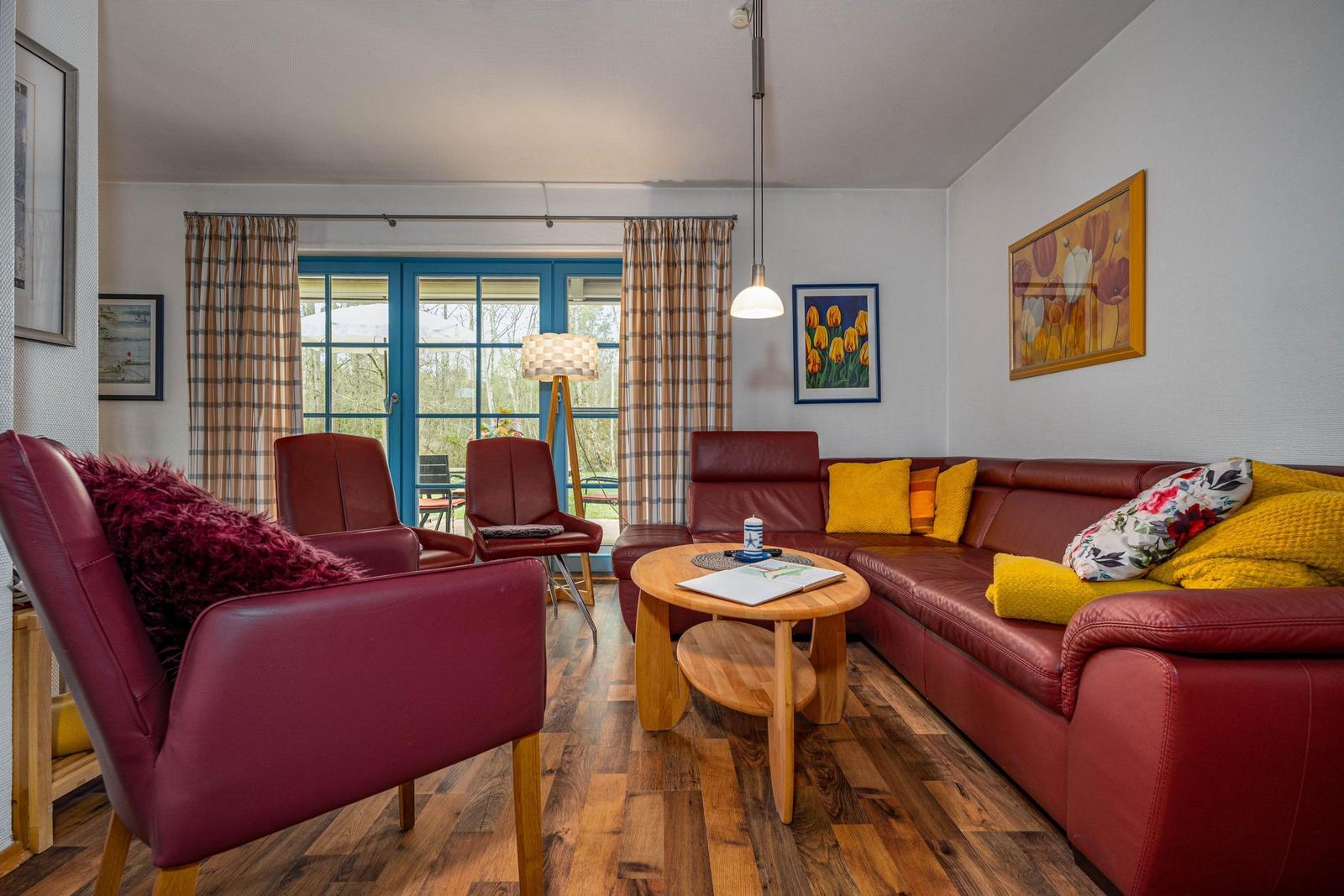 Ein Wohnzimmer mit rotem Sofa, Stühlen und Holztisch. Durch Fenster blickt man auf Garten.