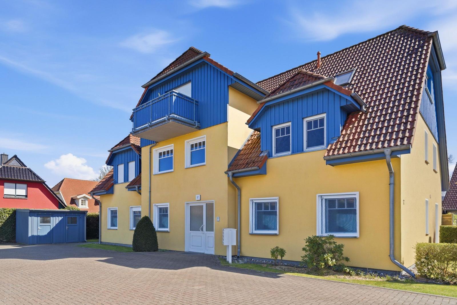 Zwei-stöckiges Haus mit gelber Fassade und blauen Dachbereichen. Vordere Tür und kleine Grünflächen.