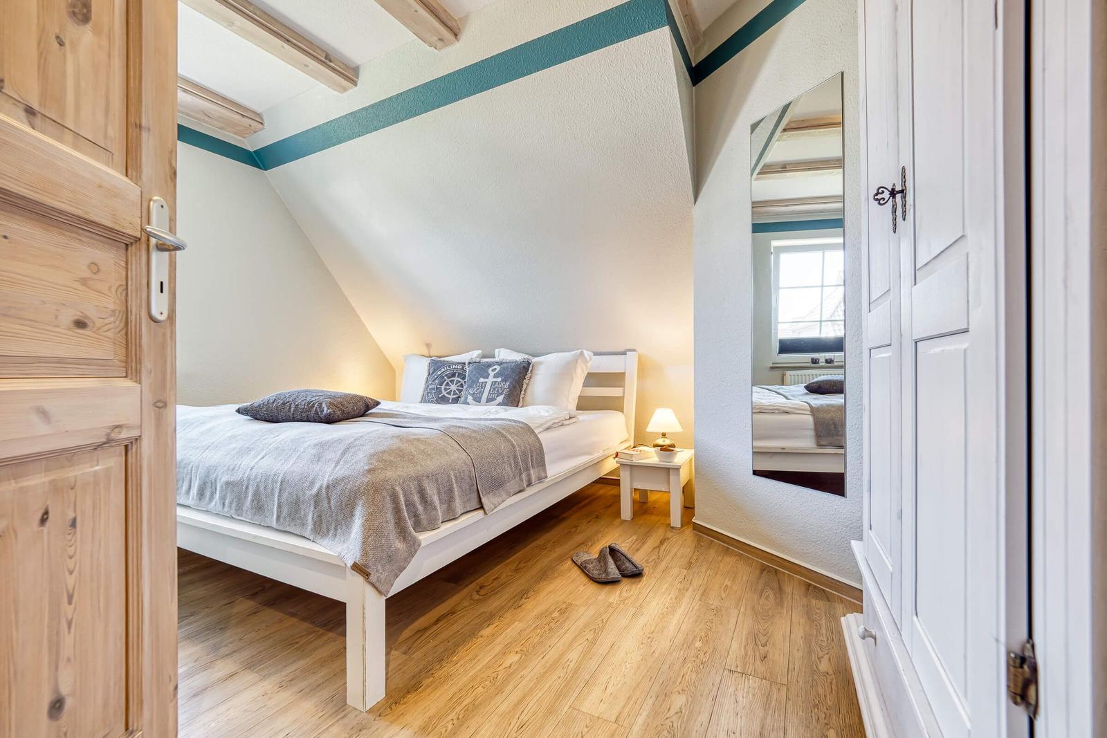Schlafzimmer mit Bett, Nachttisch und Spiegel. Holzboden und Holztür sichtbar.