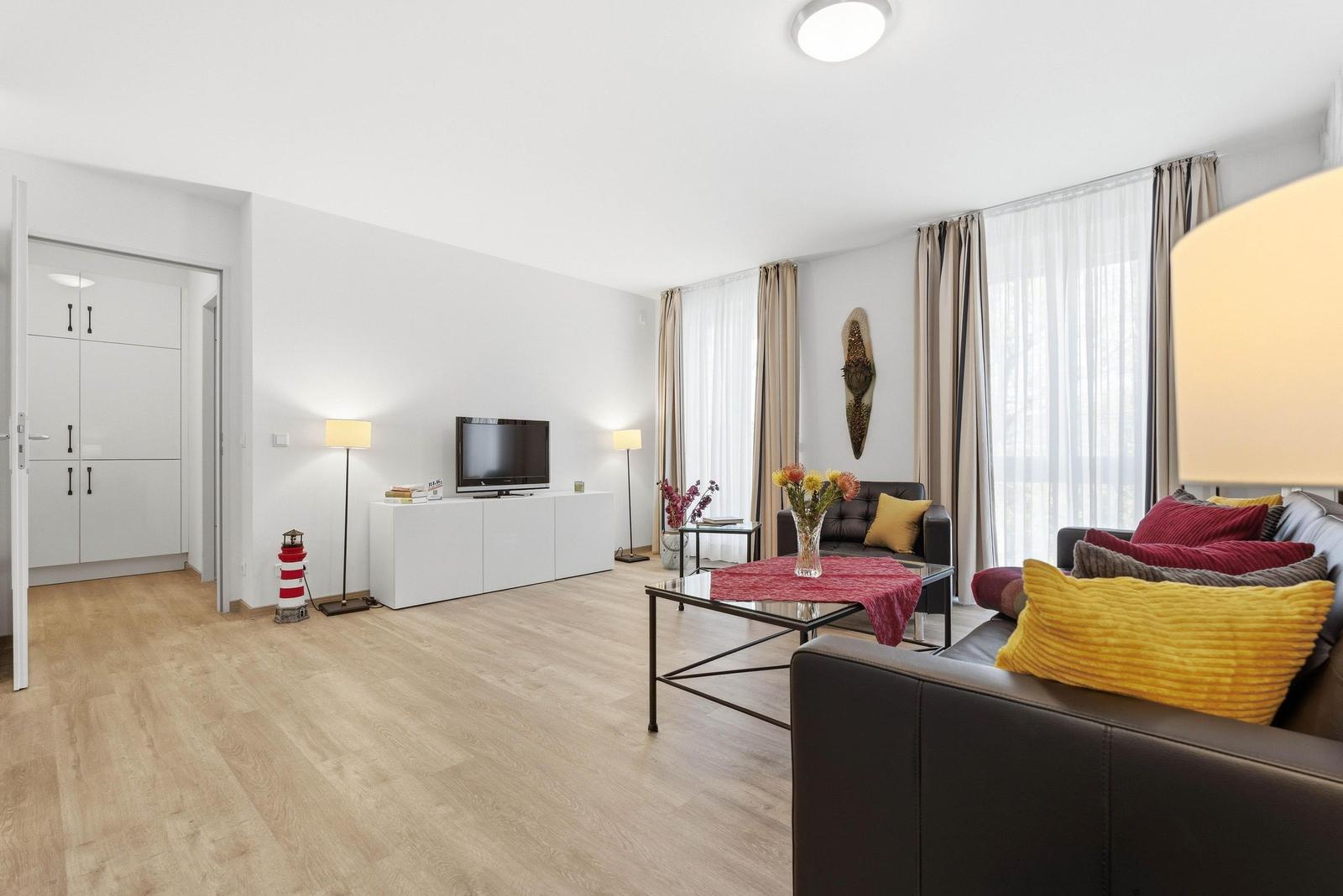 Wohnzimmer mit Sofa, Couchtisch und Fernseher. Holzboden und große Fenster.