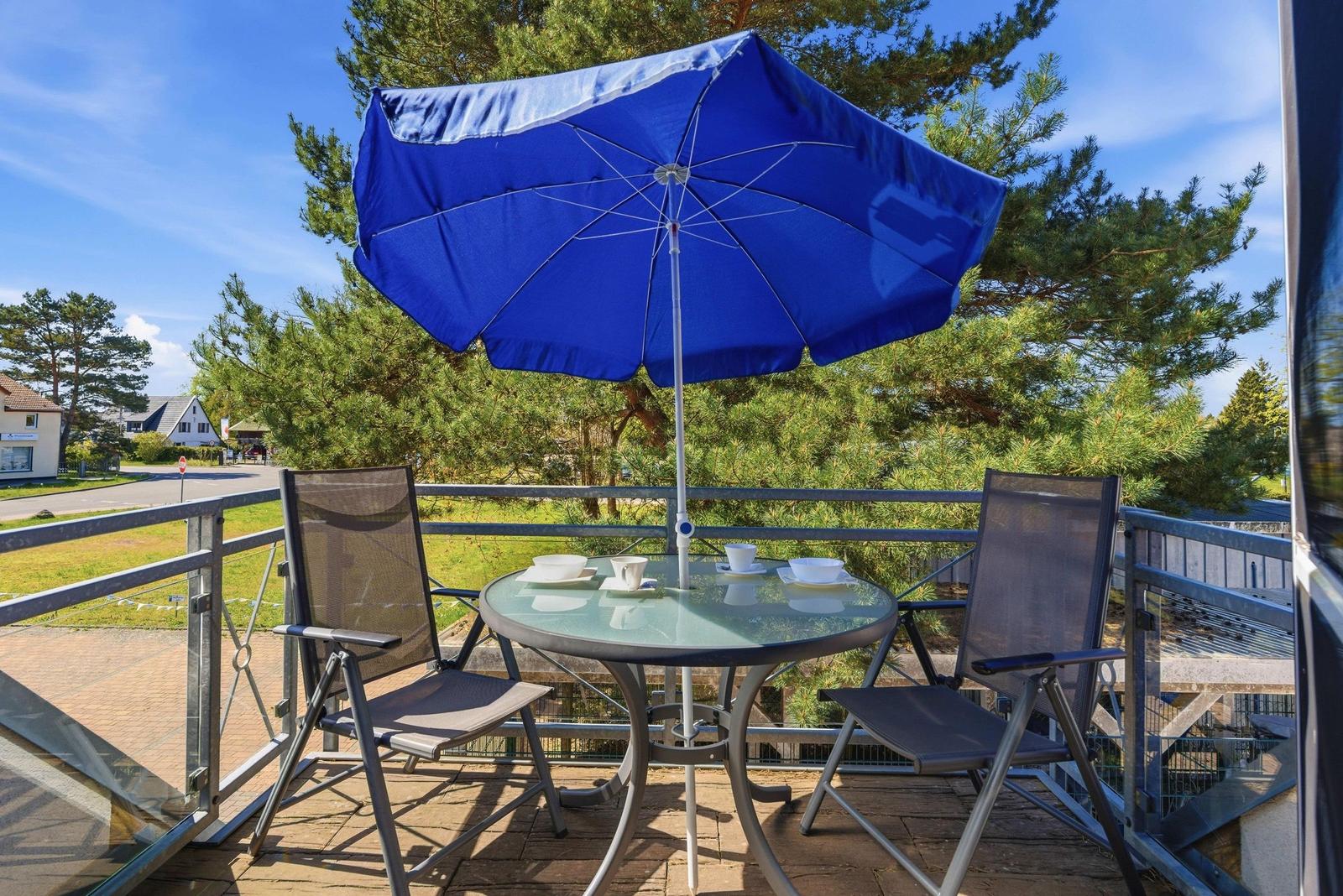 Terrasse mit Tisch, zwei Stühlen und blauem Sonnenschirm. Blick auf Bäume und Straße.
