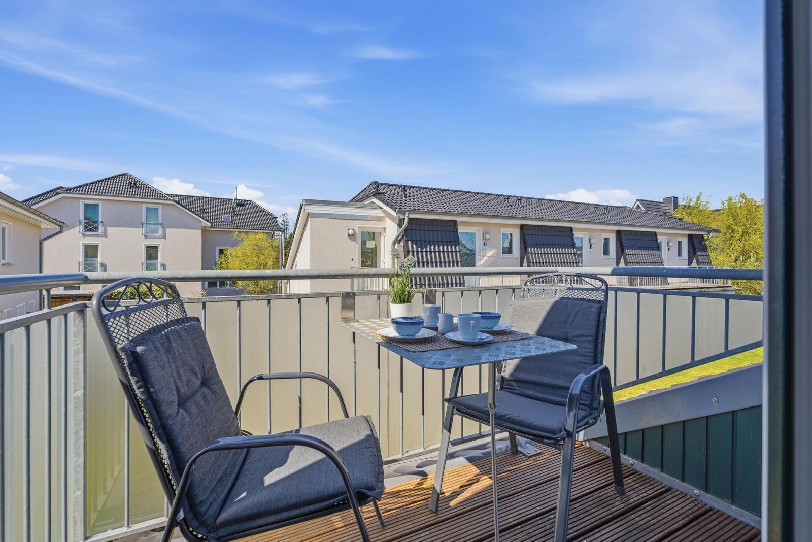 Terrasse mit Tisch und zwei Stühlen. Im Hintergrund sind Wohnhäuser und Bäume zu sehen.
