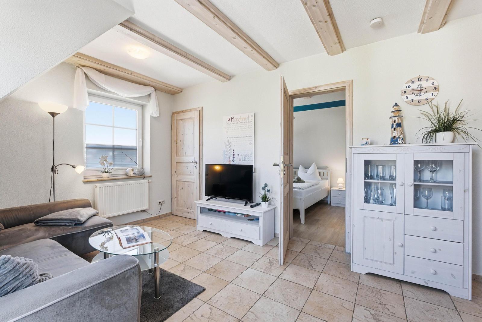Wohnzimmer mit Sofa, TV und Blick in Schlafzimmer. Holzbalken und weißer Schrank.