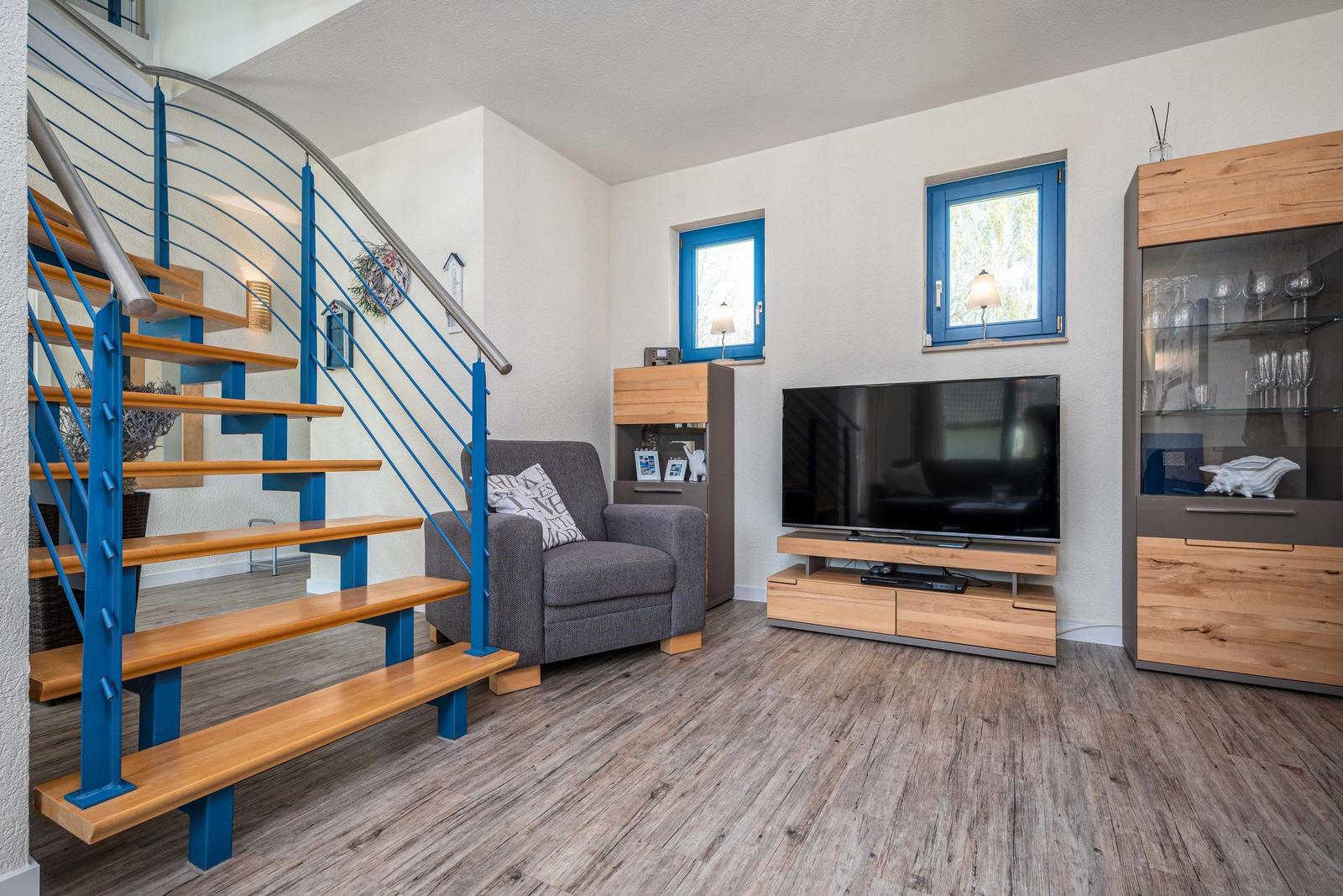 Wohnzimmer mit Fernseher, Sessel und Treppe mit blauen Metallgeländern.