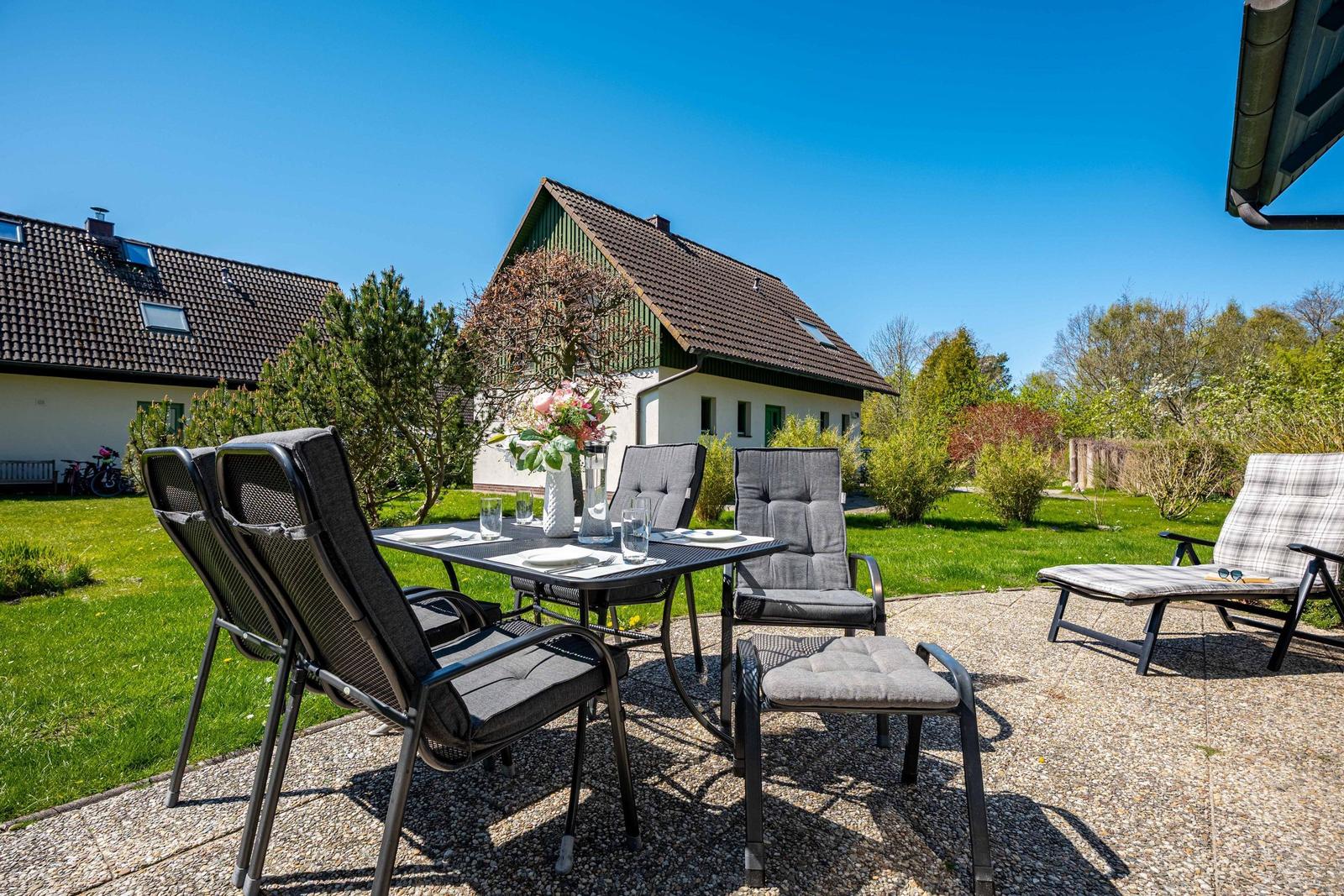 Terrasse mit Tisch, Stühlen und Liegestuhl. Hintergrund: Haus und Grünflächen.