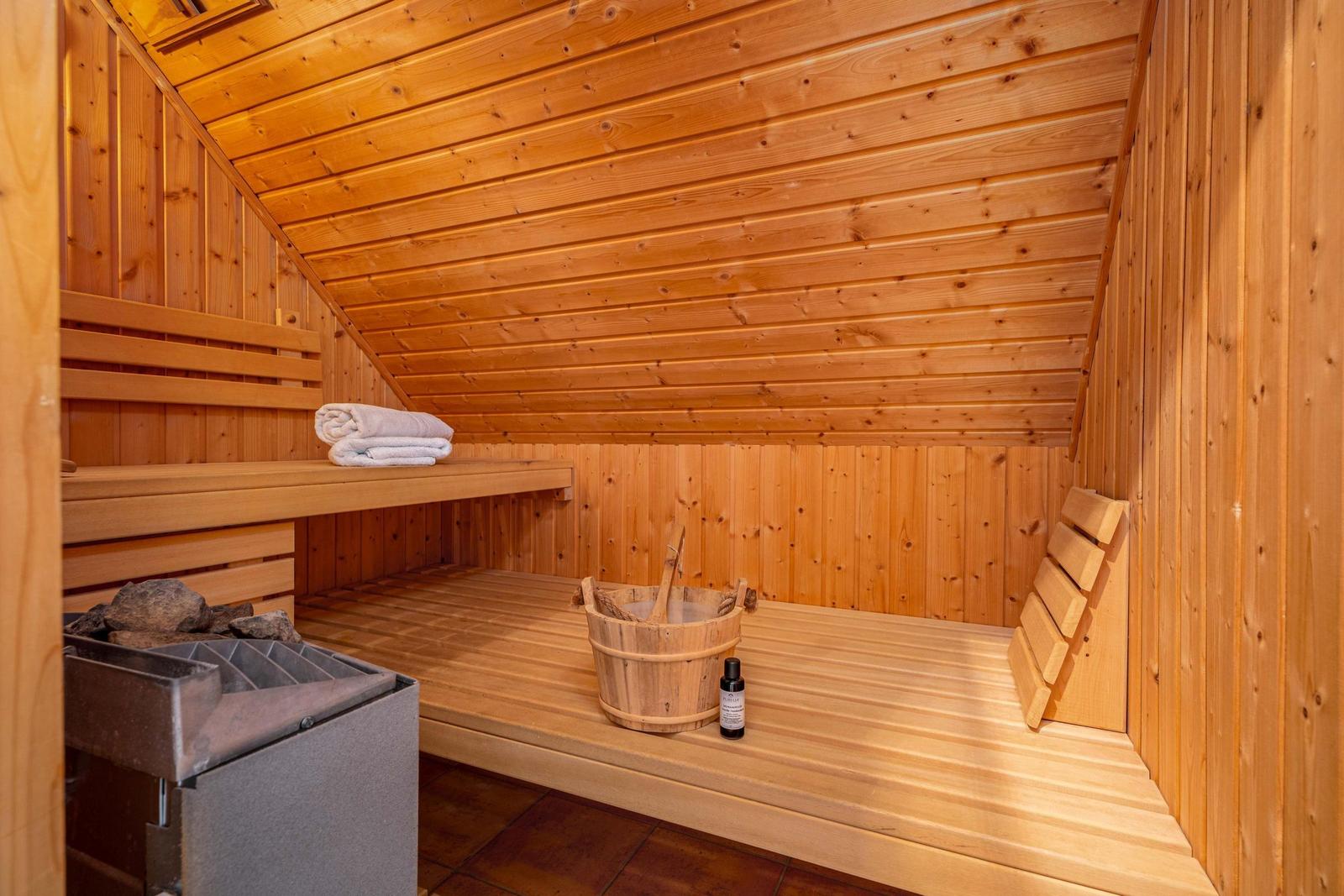 Sauna mit Holzbänken, Eimer und Handtuch. Heizung und Fliesenboden sichtbar.